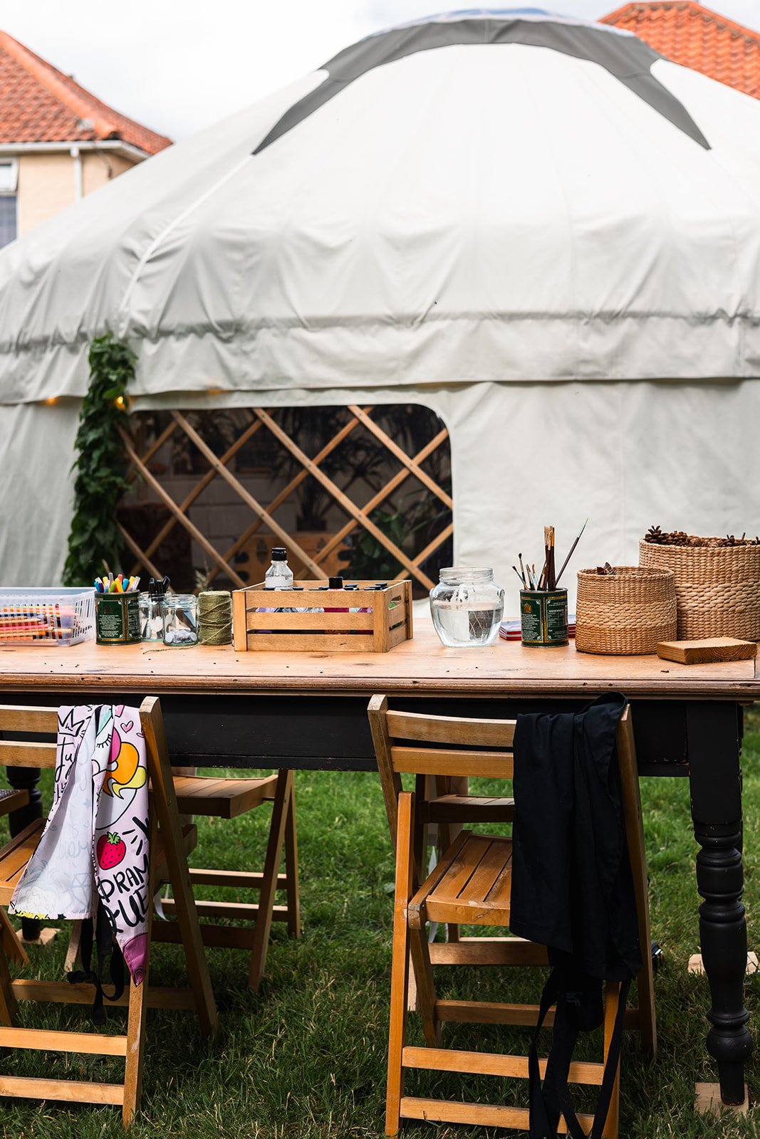 Outdoor workspace with wooden table and chairs set up outside a white canopy, with art supplies, baskets, and jars on the table.