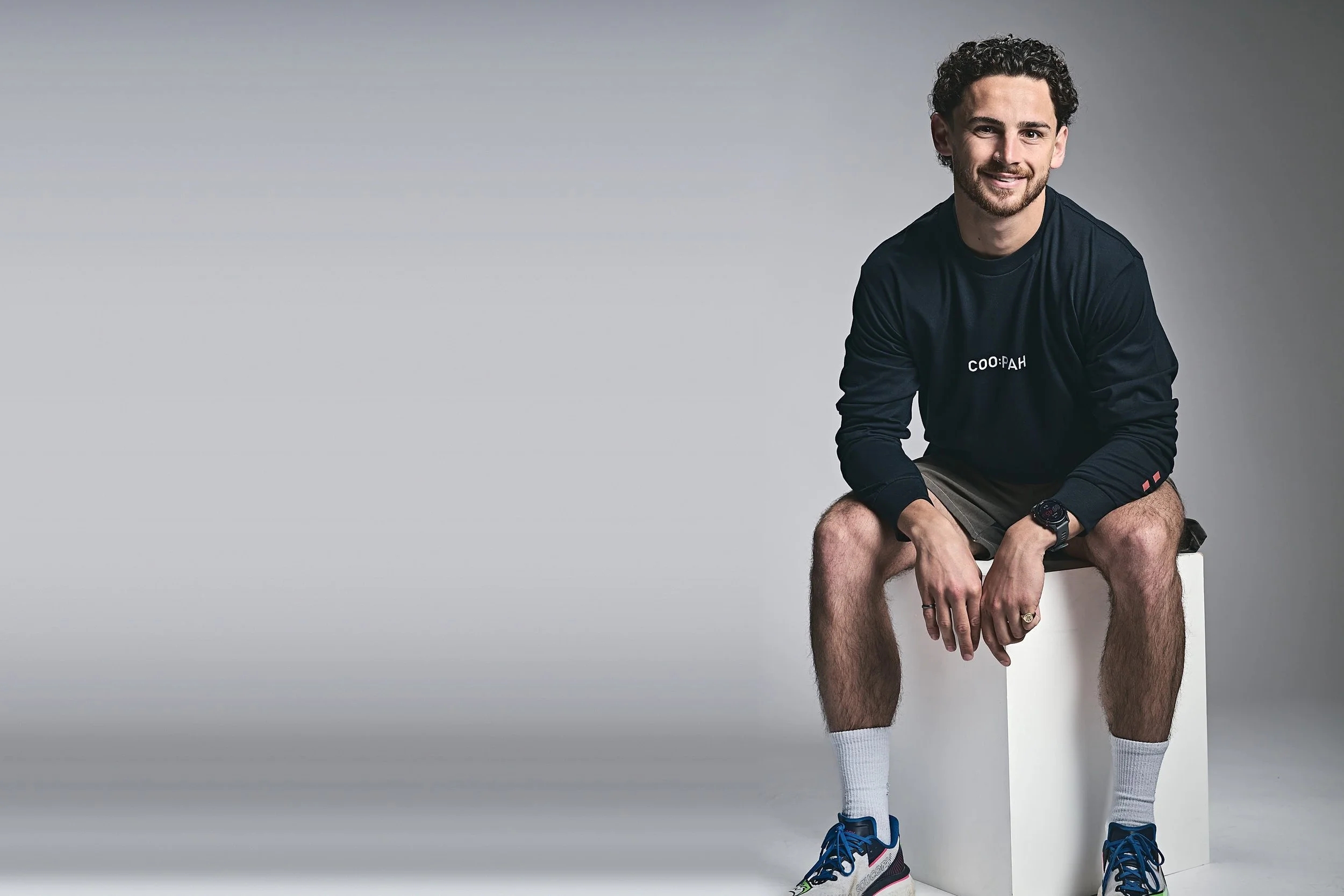 A man with dark curly hair and a beard sitting on a white block with his hands clasped in front of his knees. He is wearing a black long-sleeve shirt, gray shorts, white socks, and colorful sneakers. The background is plain gray.