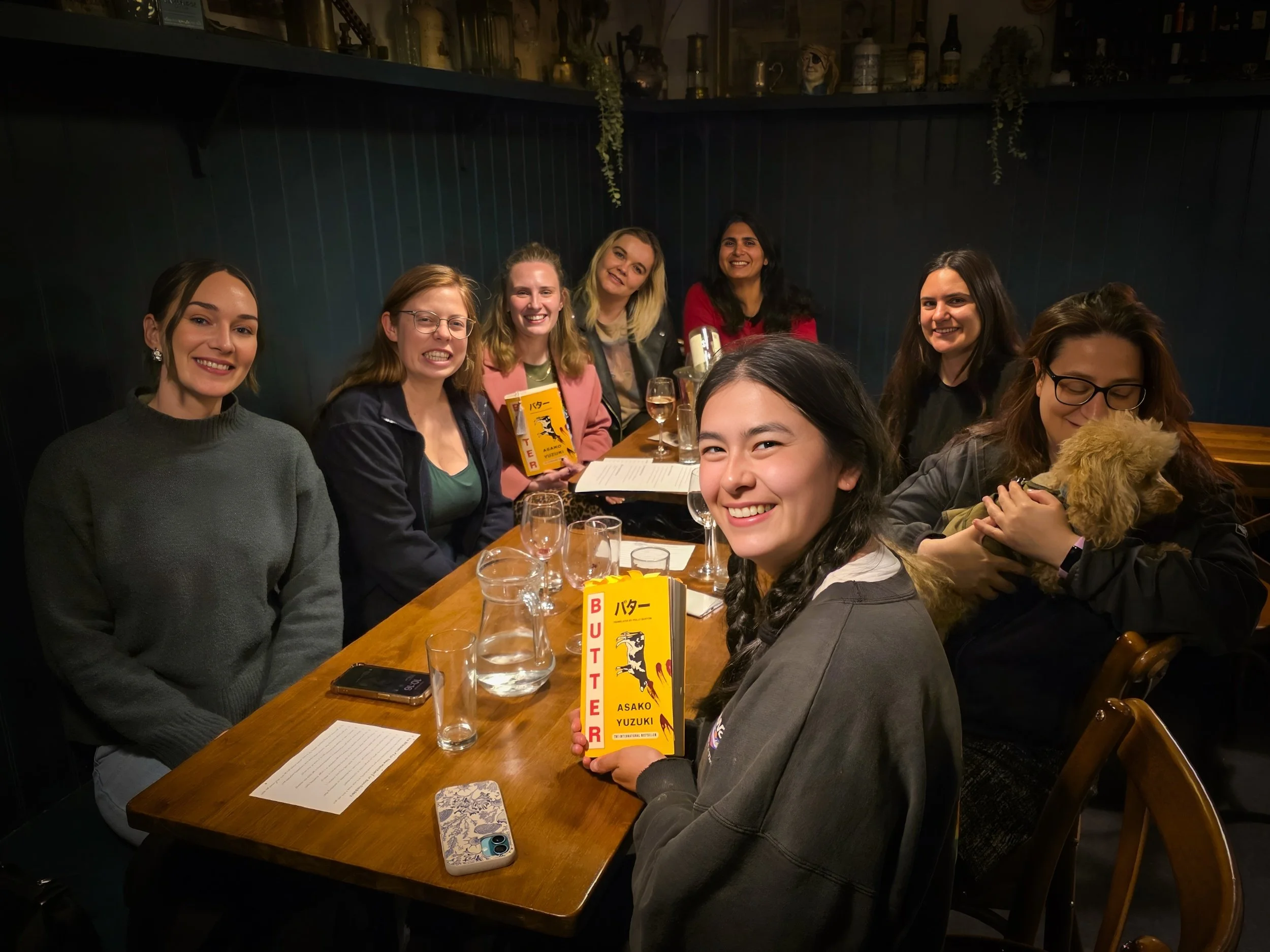 Group of nine women sitting around a wooden table in a dimly lit restaurant or bar, smiling at the camera. One woman is holding a yellow book titled 'BUTTER' by Asako Yuzuki. There are glasses, a phone, and a notepad on the table, and a woman on the right is holding a small dog.
