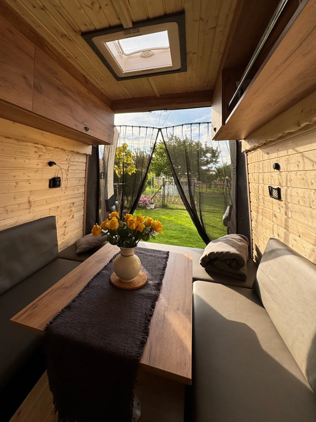Interior of a cozy camper with wooden walls and ceiling, a small skylight, and a window revealing a green backyard with trees and a garden. There is a wooden table with a black runner and a vase of yellow roses, surrounded by cushioned benches.