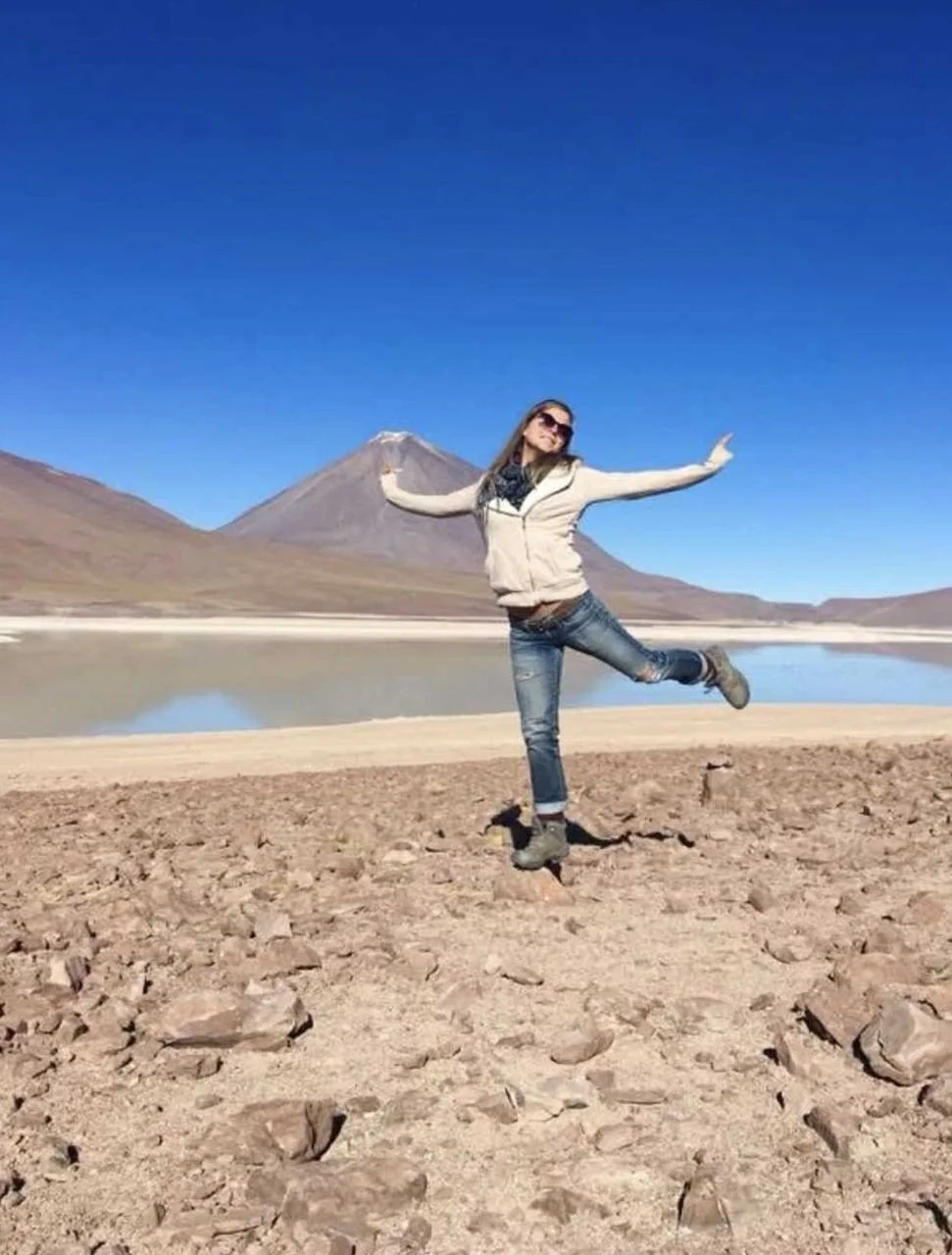 Une femme en jeans et veste, heureuse, semblant sauter en l'air sur un terrain rocheux avec un lac et une montagne en arrière-plan, avec un ciel bleu clair parfait.