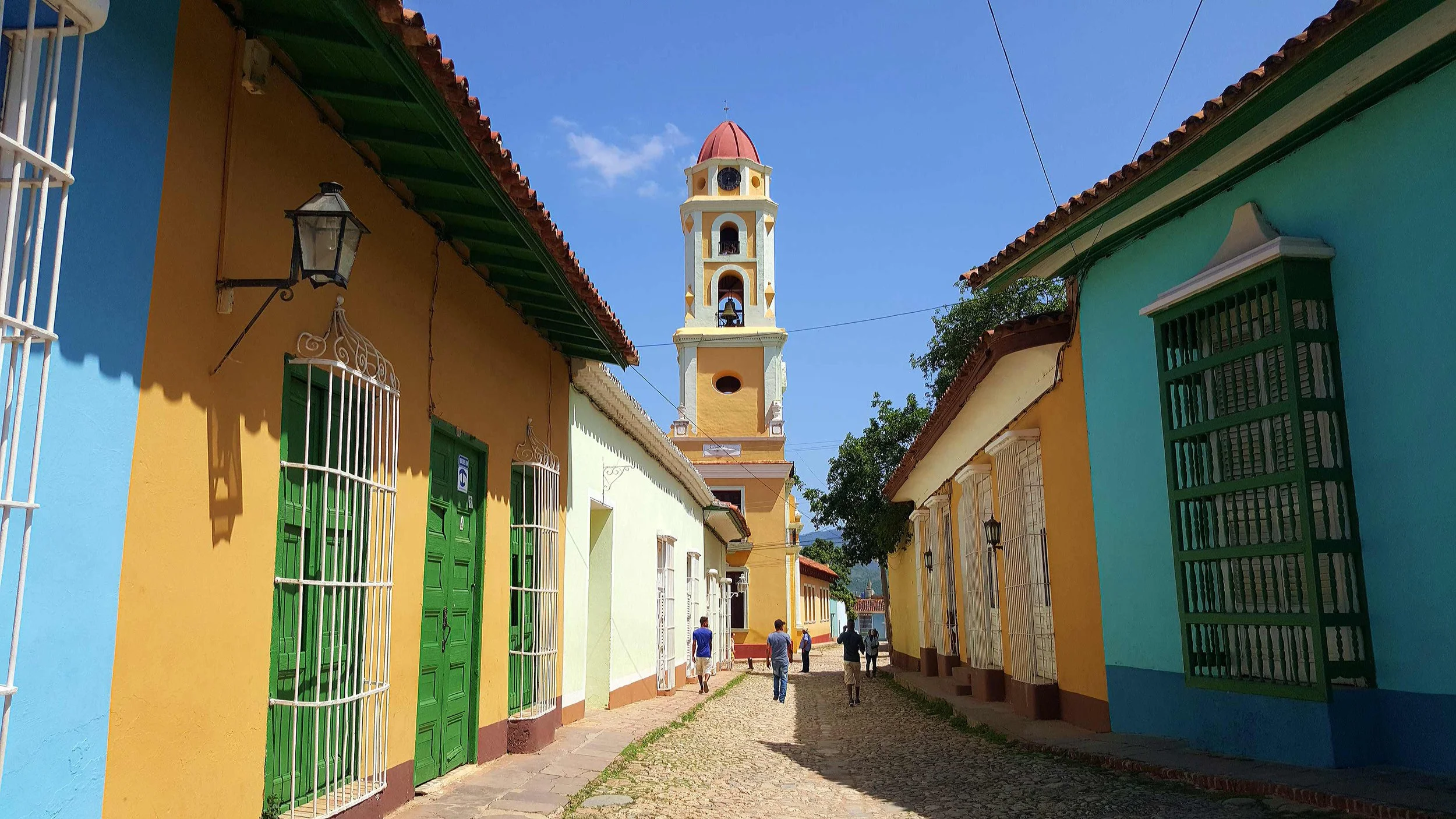 Rue pavée de Trinidad avec des maisons colorées en jaune, vert et bleu, avec des barreaux de sécurité aux fenêtres, et une église blanche avec un clocher jaune et rouge au centre.