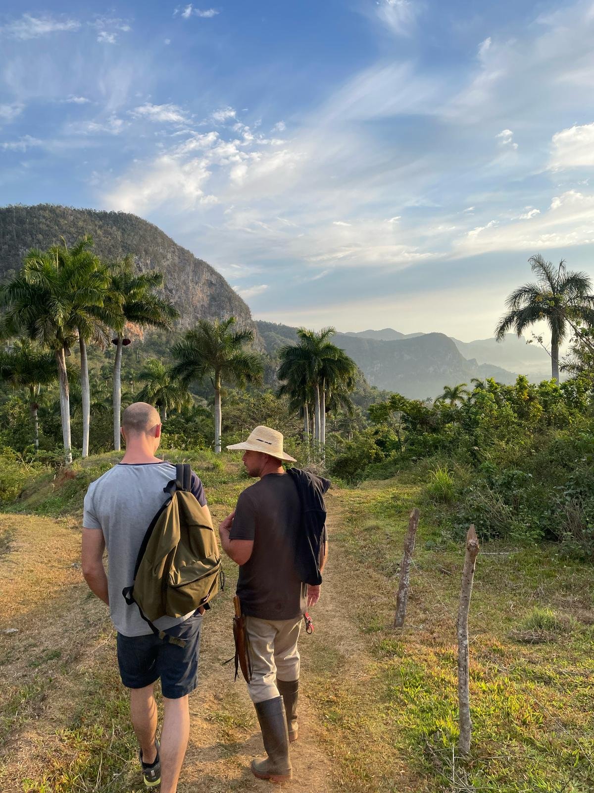Deux personnes marchant sur un sentier dans la vallée de Vinales avec des palmiers et des montagnes en arrière-plan, sous un ciel partiellement nuageux.