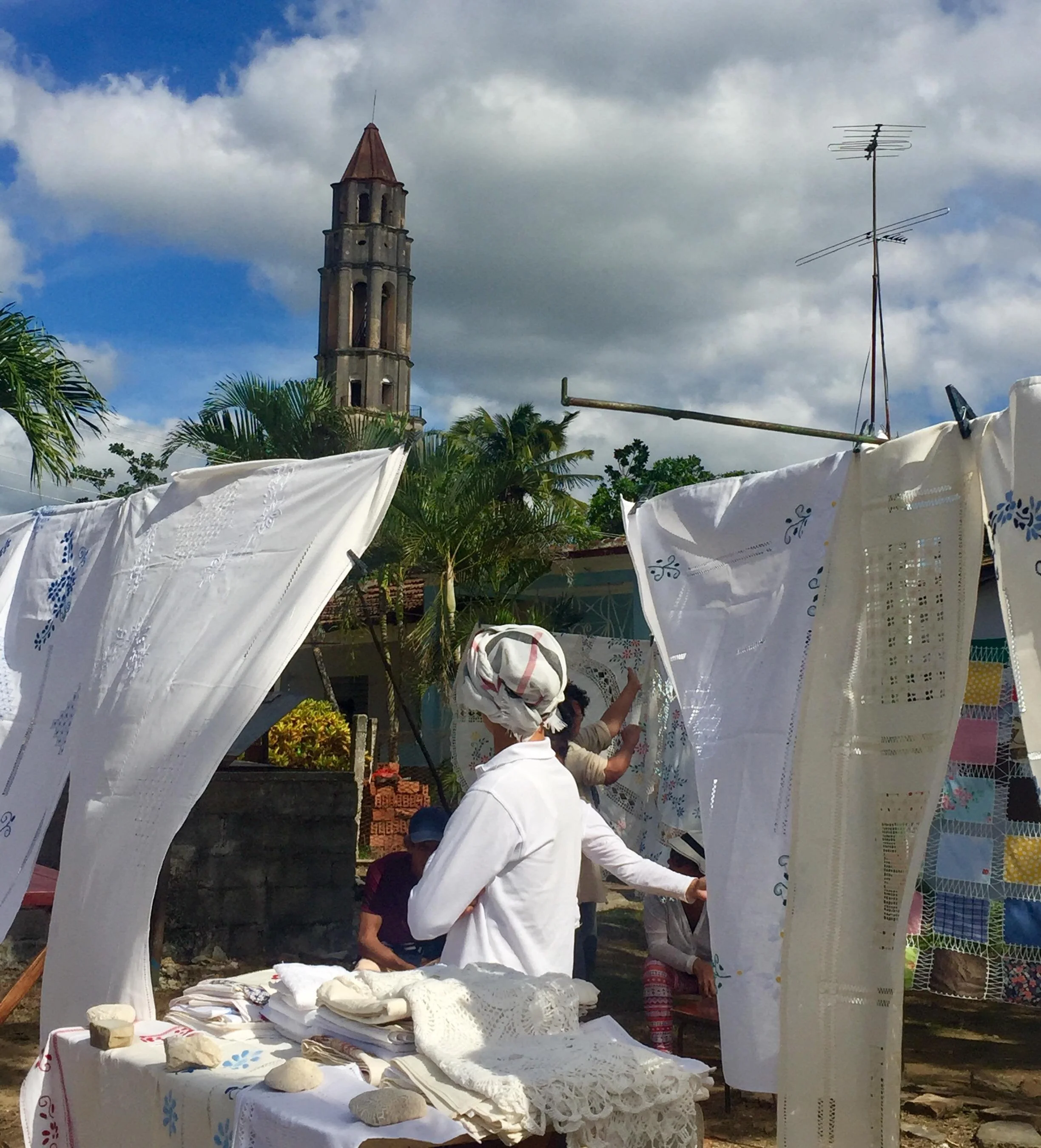 Marché en plein air à Trinidad avec des tissus brodés et des personnes discutant, la Tour Iznaga en pierre en arrière-plan.