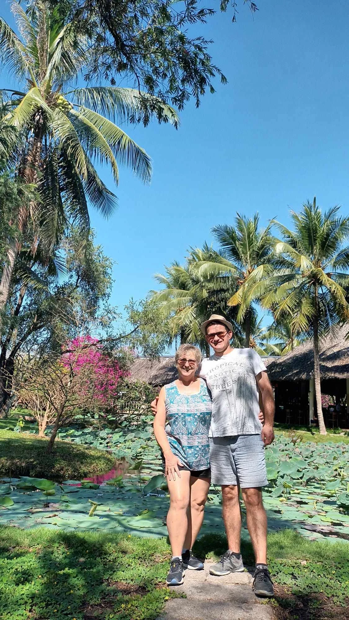 Nos deux voyageurs souriants posent ensemble dans un jardin tropical vietnamien avec des palmiers, un étang avec des nénuphars, et une maison avec un toit en paille, sous un ciel bleu.