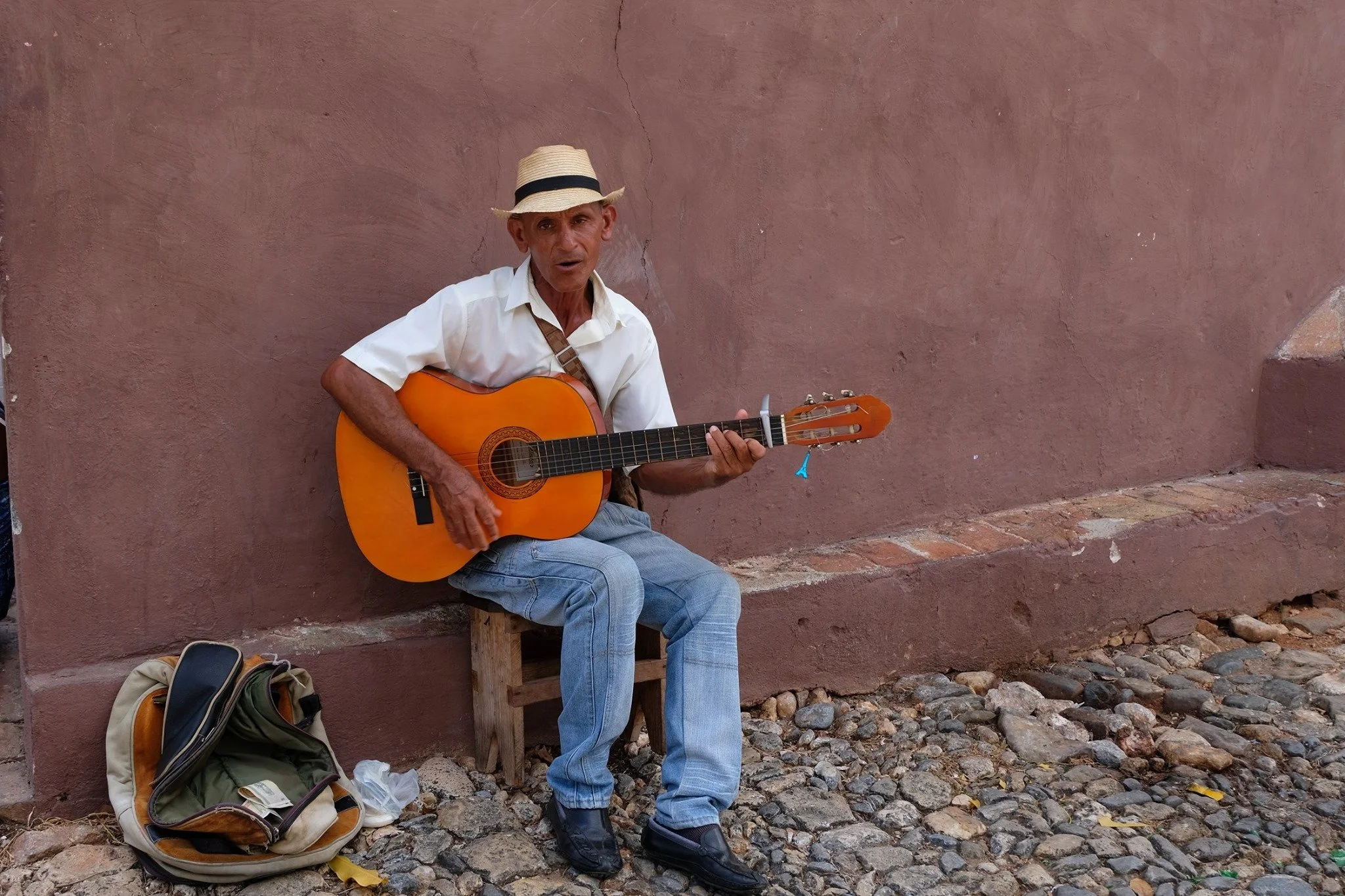 Un homme âgé jouant de la guitare assis sur un petit tabouret devant un mur en pierre rose