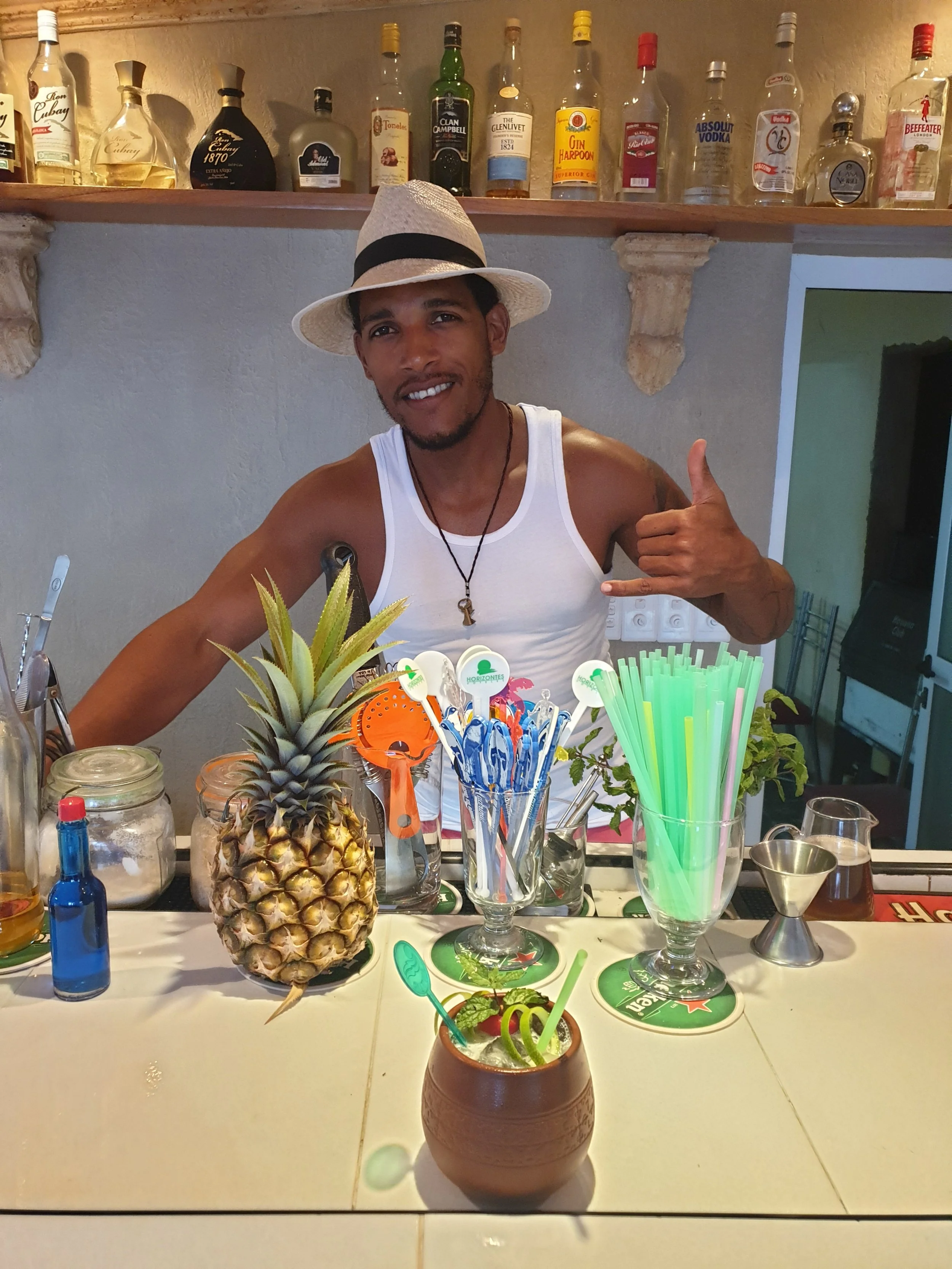 Un barman cubain souriant portant un chapeau de paille blanc, debout derrière son bar avec devant lui 2 délicieux cocktals qu'il vient de réaliser.