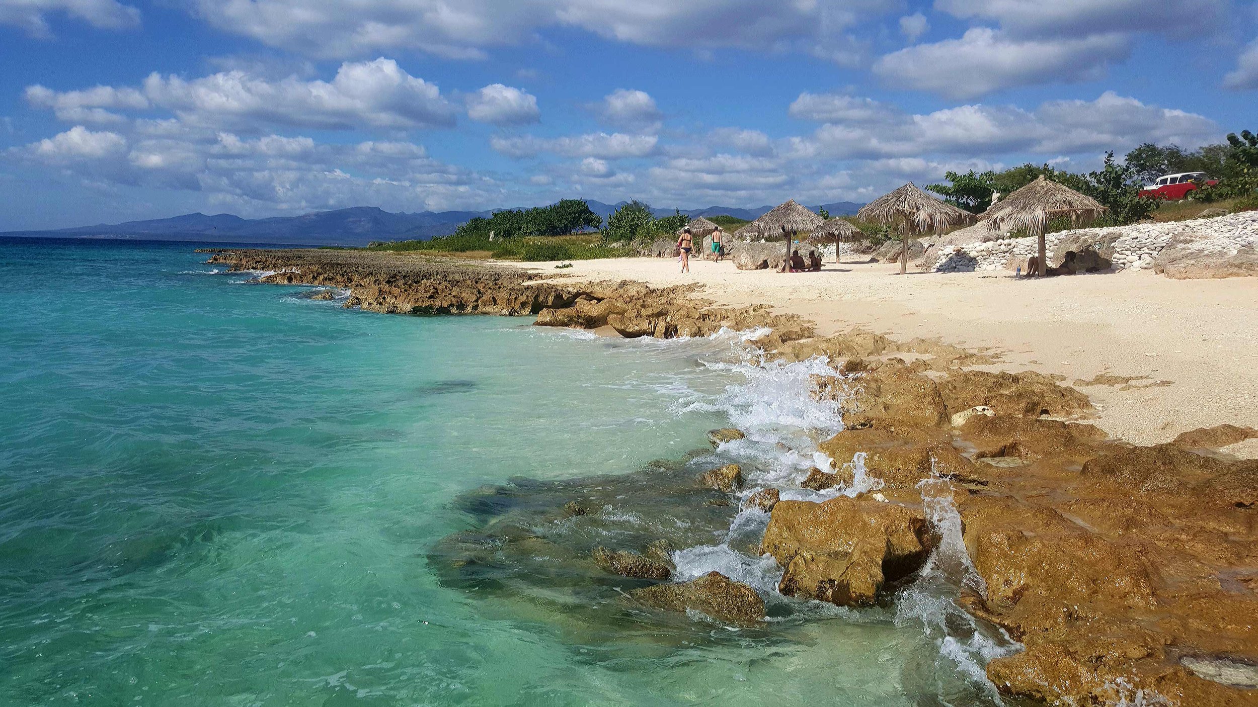 Plage de sable blanc à Trinidad avec des parasols en paille, quelques rochers dans l'eau turquoise, et les montagnes de l'Escambray au loin.