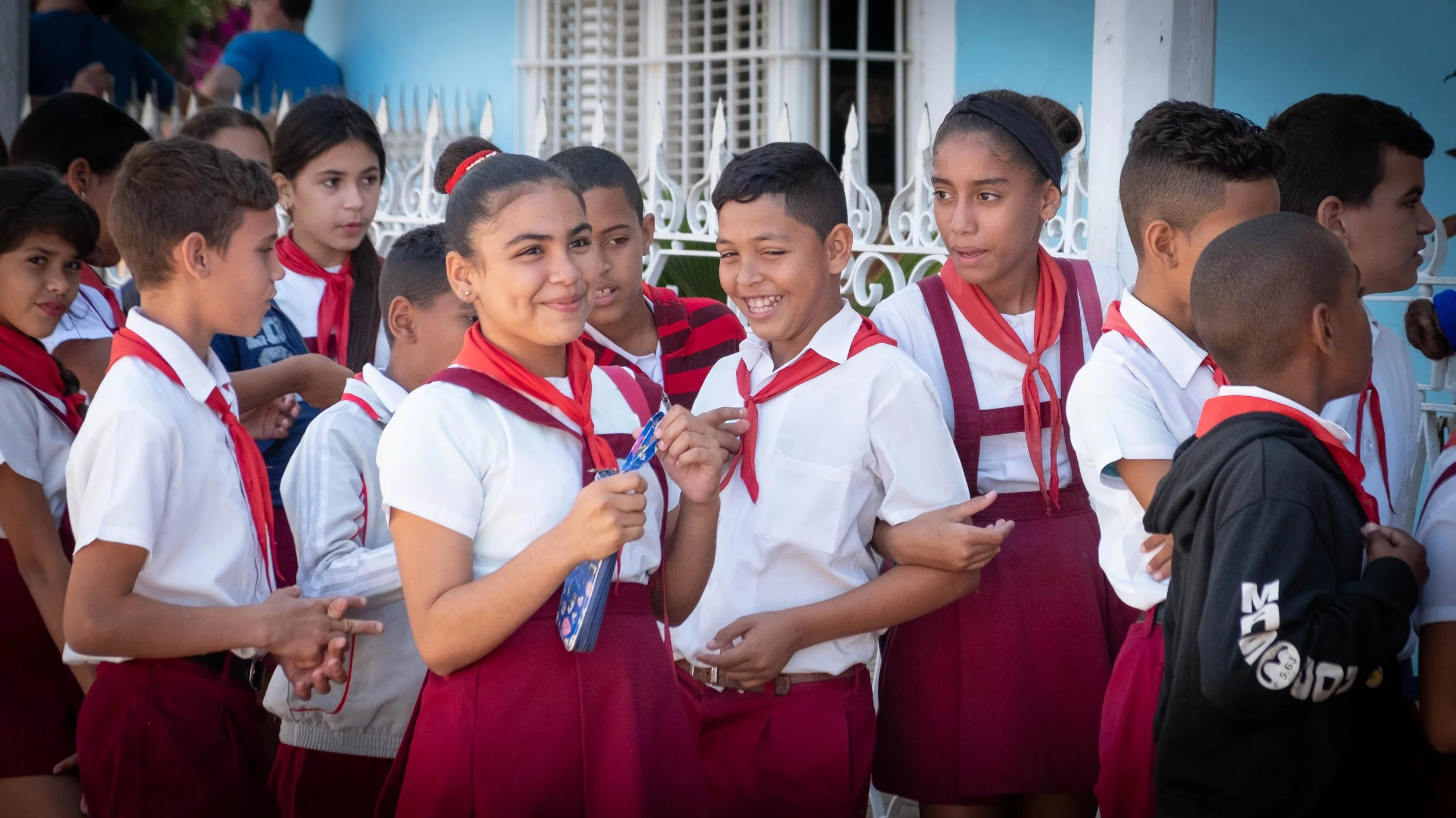 Groupe d'élèves cubains en uniforme scolaire rouge et blanc, devant une grille blanche et un bâtiment bleu, en ligne ou en groupe, fiers et heureux.