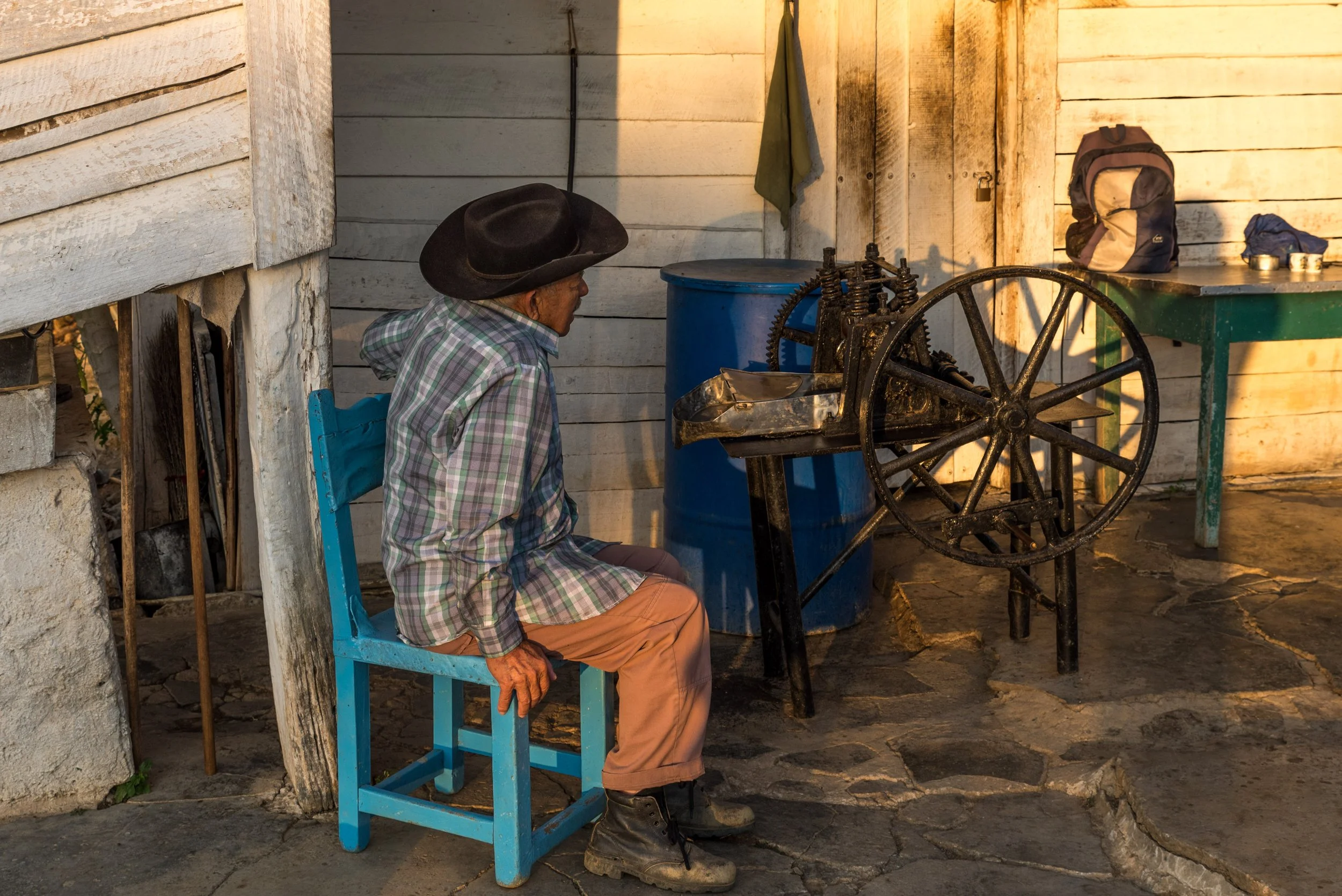 Un vieil homme assis sur une chaise bleue à côté d'une machine en fer dans une pièce en bois, avec un sac à dos sur une table à droite.