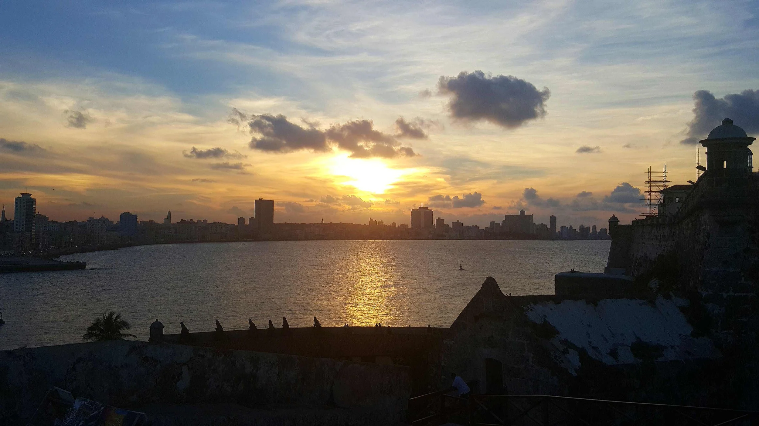 Coucher de soleil sur la ville avec des bâtiments modernes en silhouette, vue sur l'océan avec quelques nuages dans le ciel