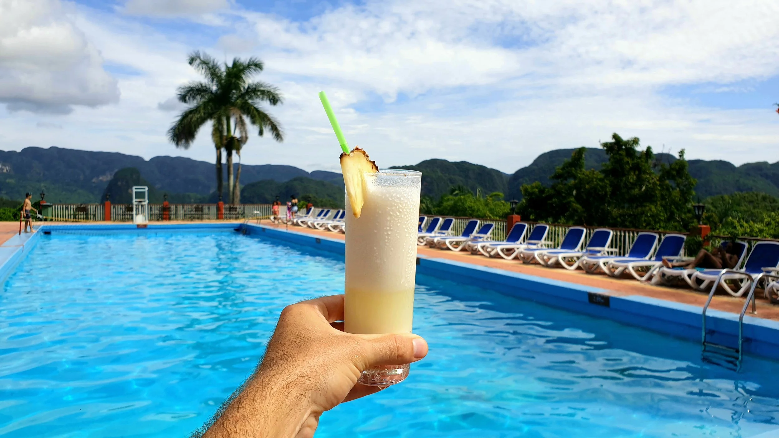 Main tenant une pina colada devant une piscine avec des transats et des palmiers en arrière-plan.
