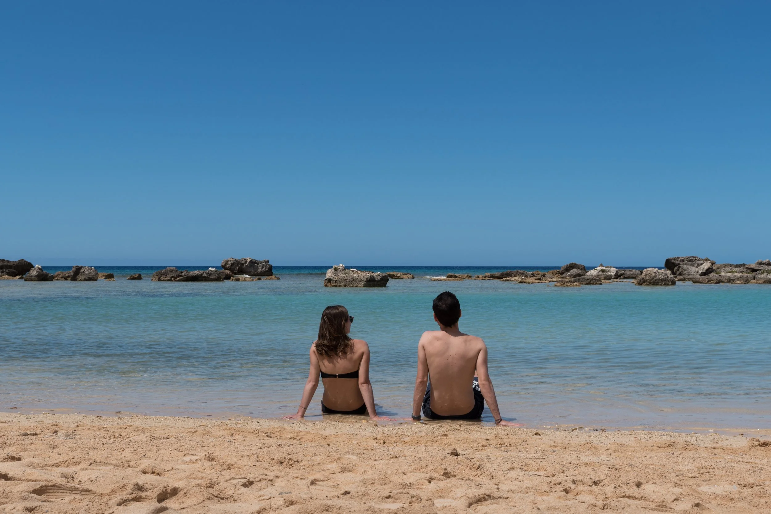 Deux personnes assises sur la plage regardant la mer, avec des rochers au loin et un ciel bleu clair.