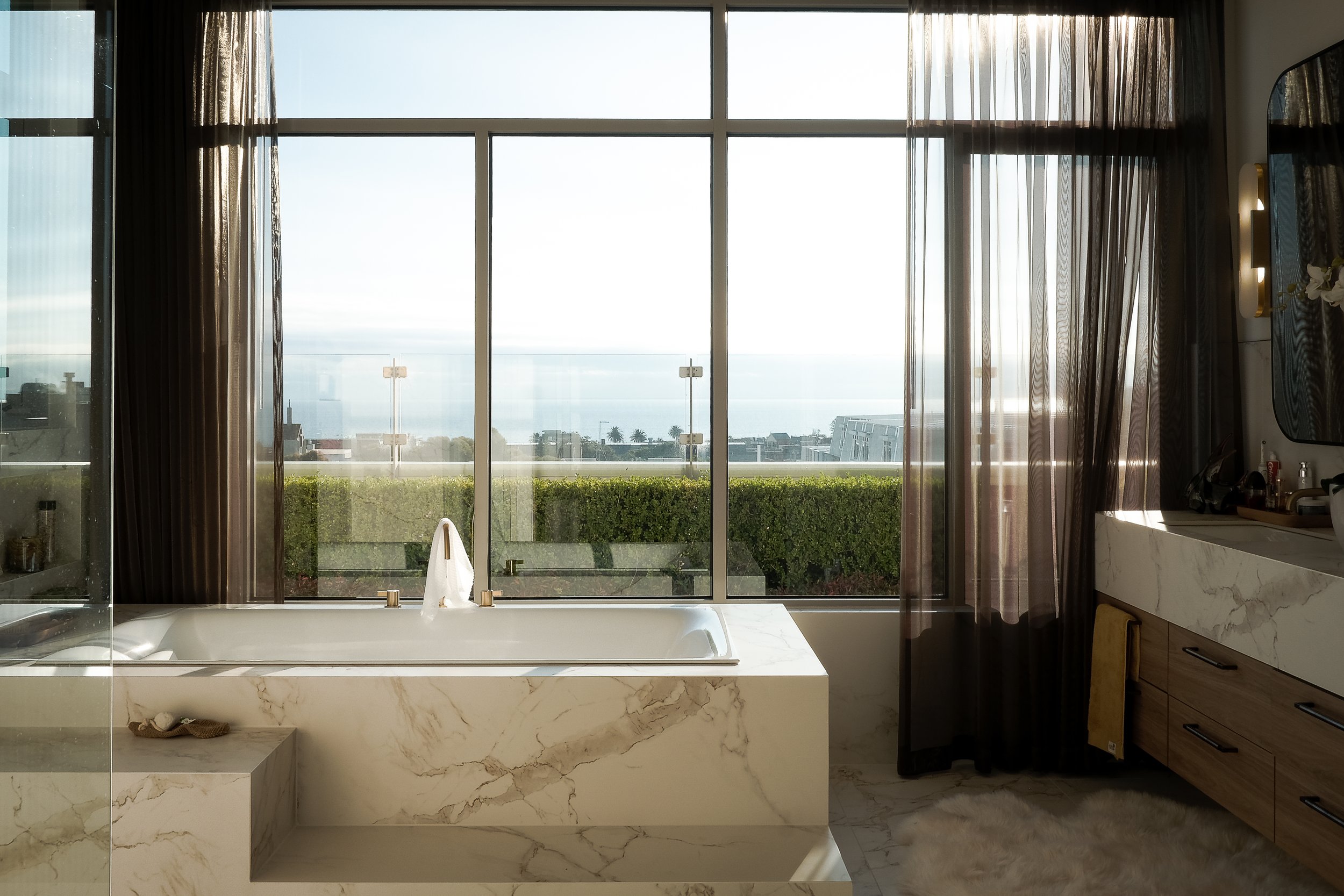 Modern bathroom with marble countertops, dual sinks, large mirrors, and a bathtub, illuminated by natural light and wall-mounted lights.