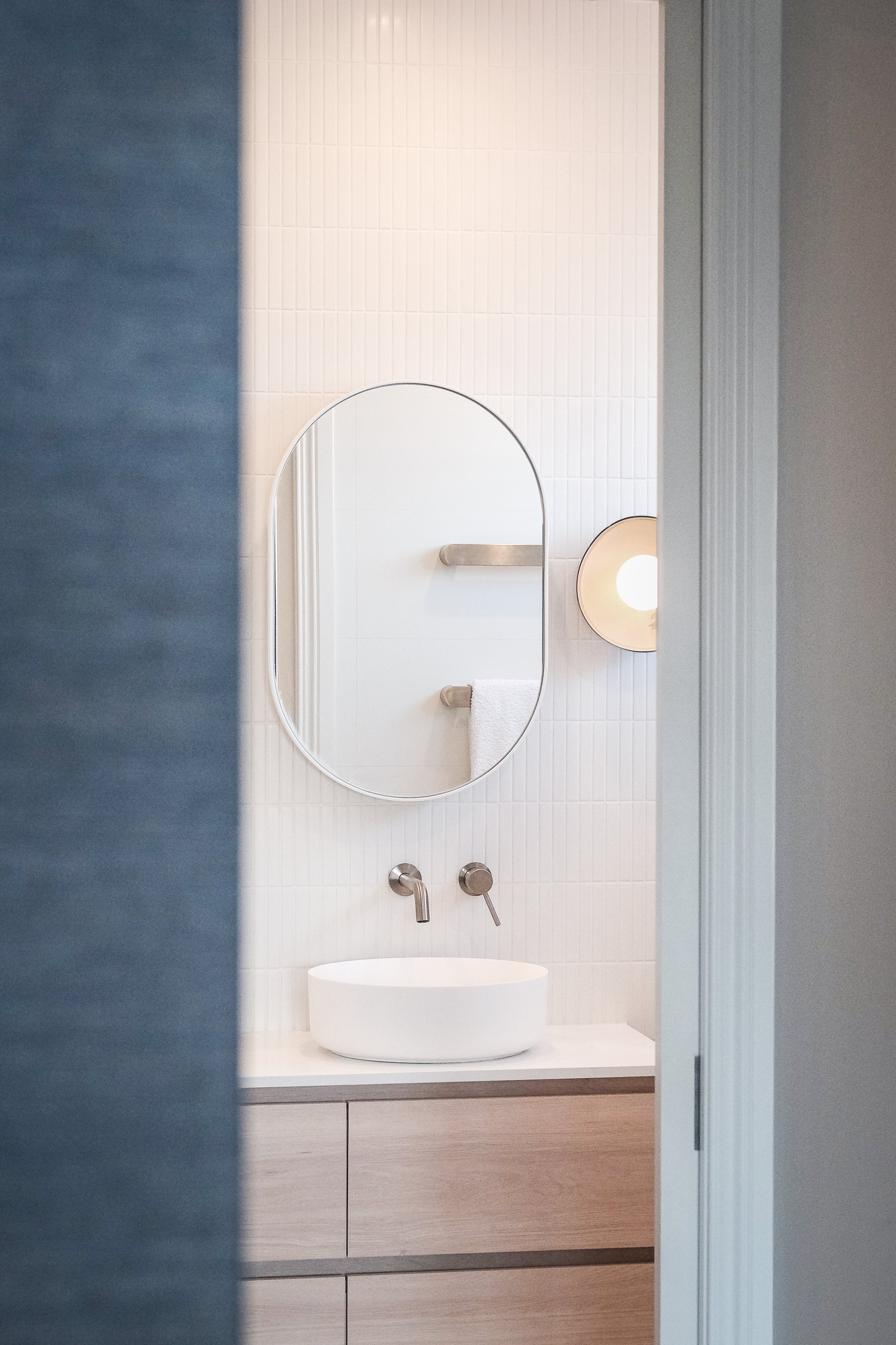 Modern bathroom with a white vessel sink, oval mirror, wall-mounted faucet, round light fixture, white tile wall, towel rack with a towel, and a small basket with white flowers.