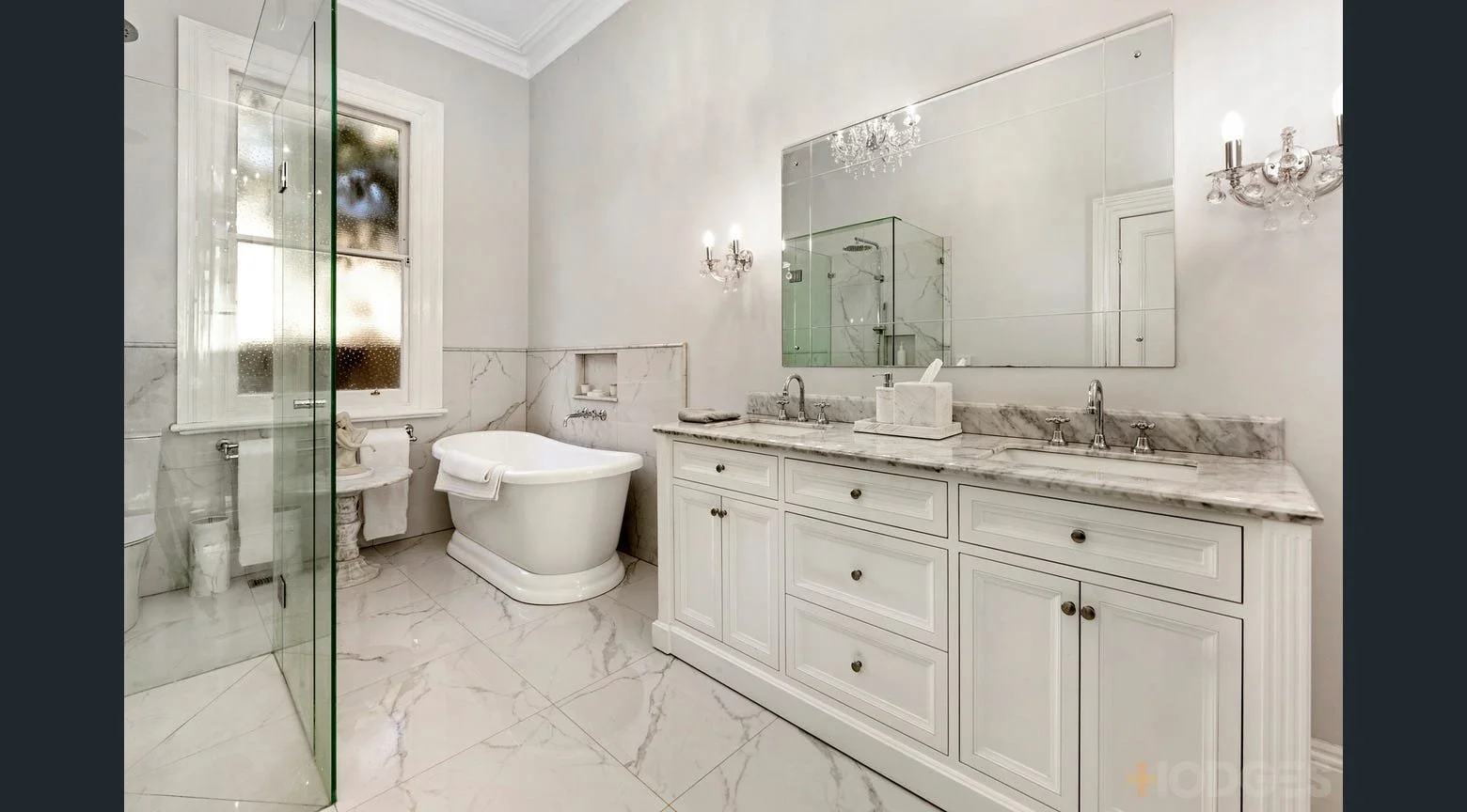 Elegant bathroom with dual sinks on a marble countertop, a large mirrored wall, crystal chandeliers, a white bathtub, vanity, and a window with a views of trees.