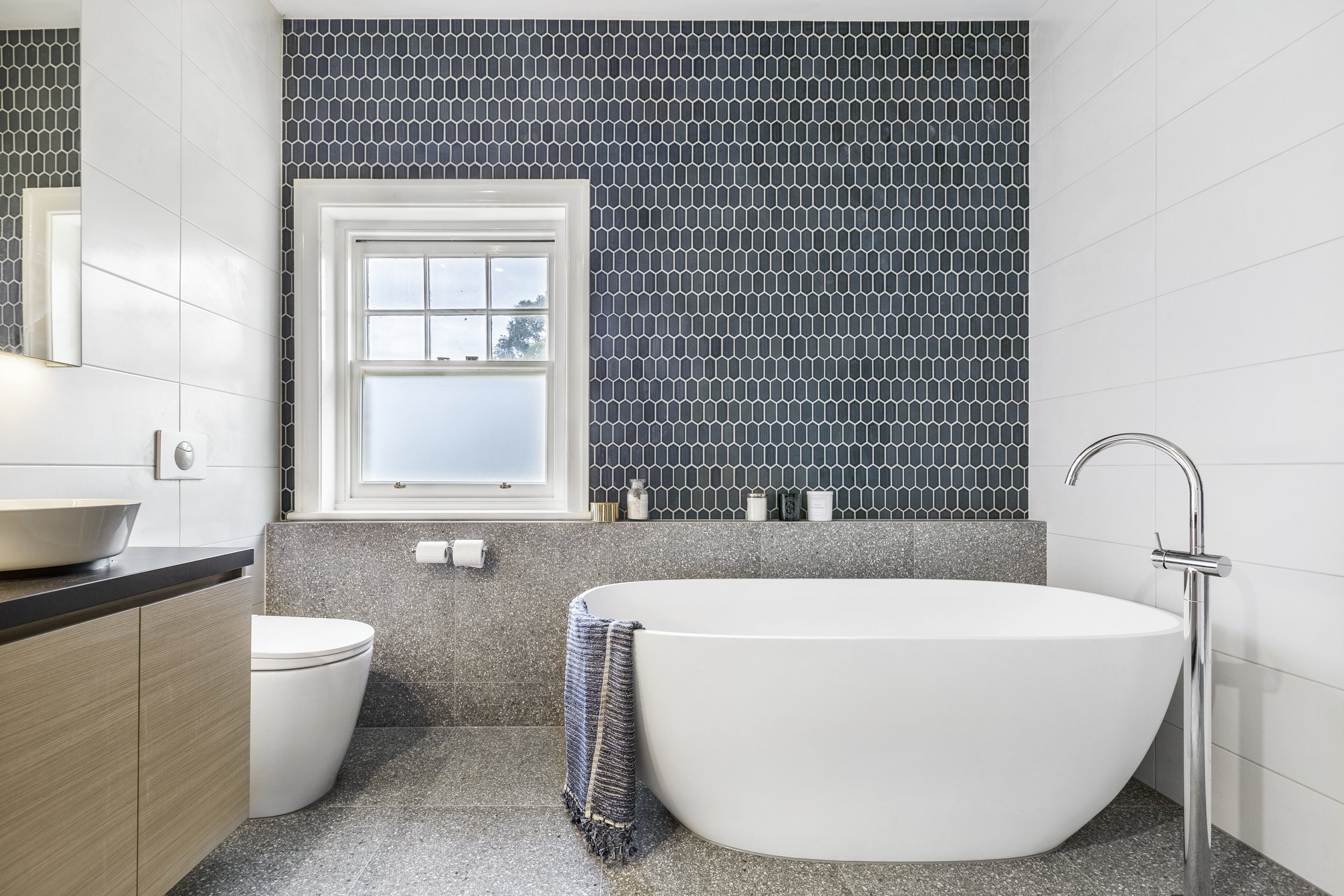 Modern bathroom with a white freestanding bathtub next to a curved wall with dark hexagonal tiles and a towel rack with blue and white towels.