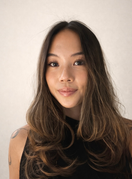 A young woman with long, wavy hair smiling with one eye slightly closed, wearing a black sleeveless top against a plain background.