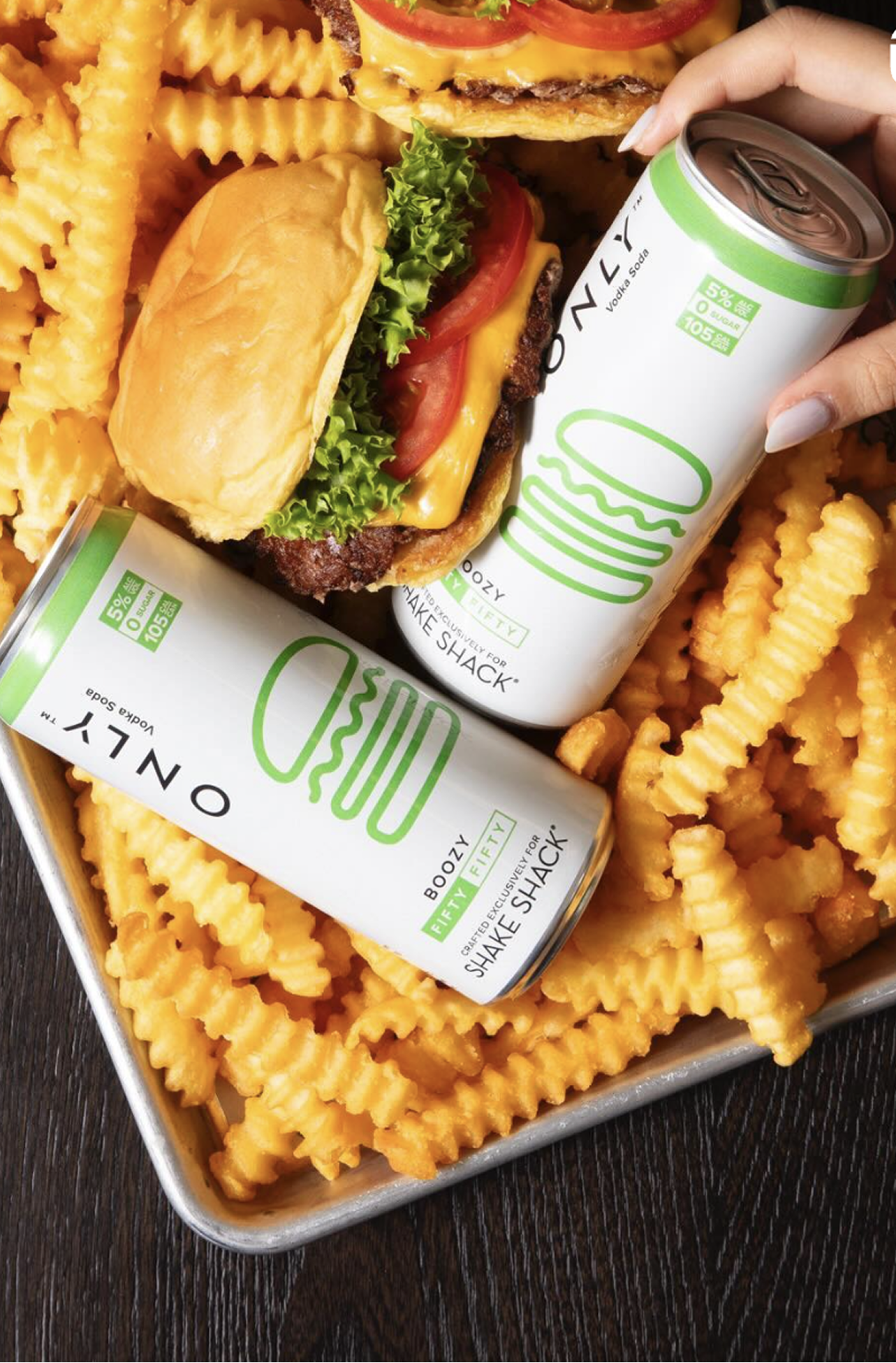 Frosted waffle fries surrounding a cheeseburger with lettuce, tomato, cheese, and a beef patty, along with two cans of Boozy Paddle, a vodka soda beverage, on a metal tray.