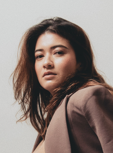 A woman with shoulder-length brown hair looking at the camera, wearing a brown blazer against a plain light background.
