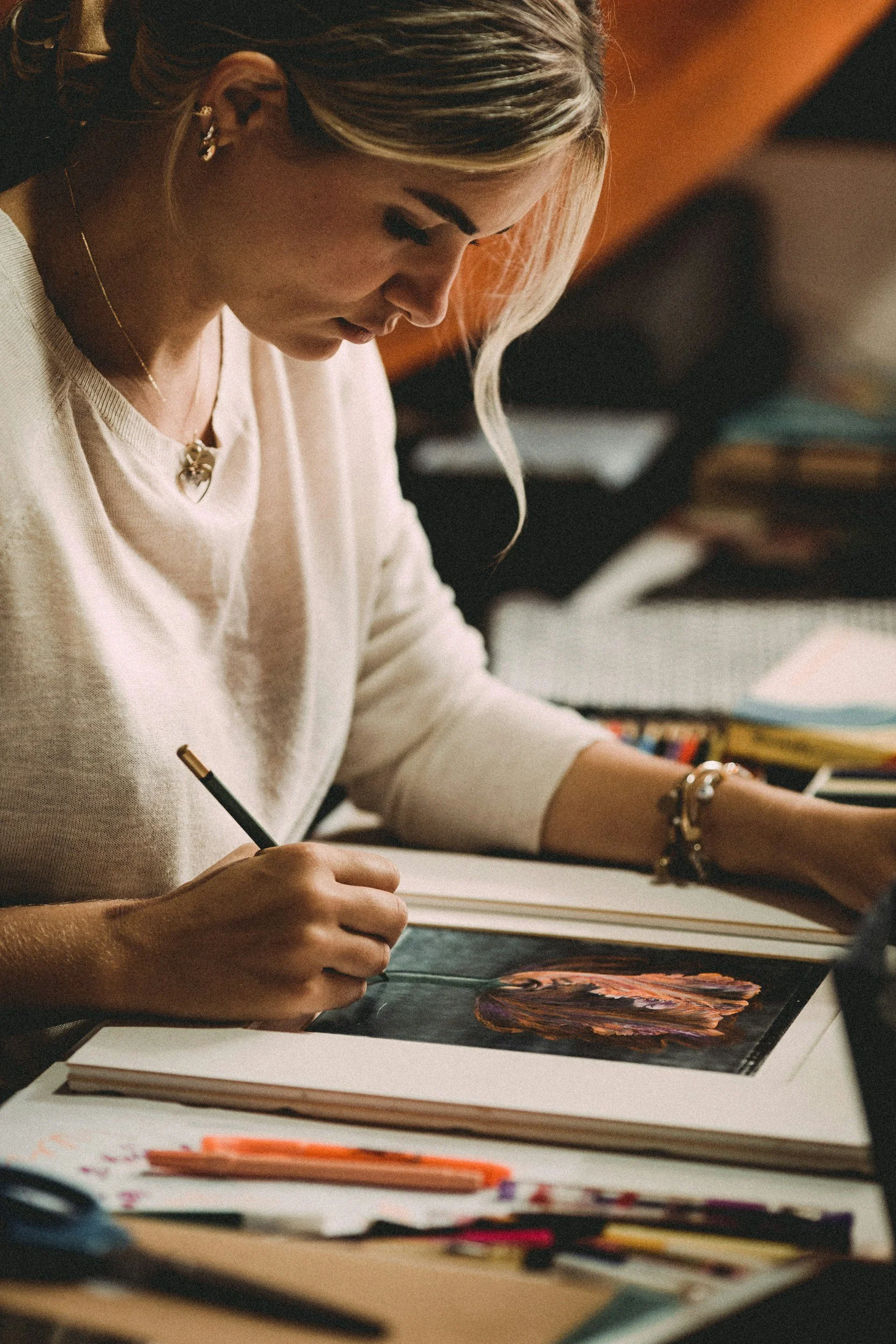 Cedar Coaching - A woman is drawing a colorful portrait of a woman with long hair on a sketchbook using colored pencils, sitting at a desk surrounded by art supplies.