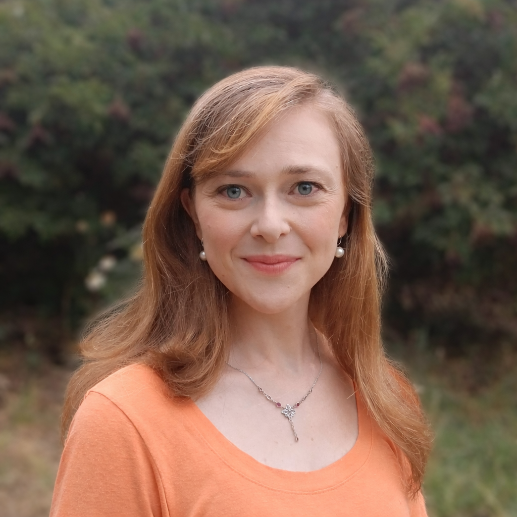 A woman with long red hair, blue eyes, wearing pearl earrings, a necklace with a bow pendant, and an orange top, standing outdoors with a blurred background of green foliage.