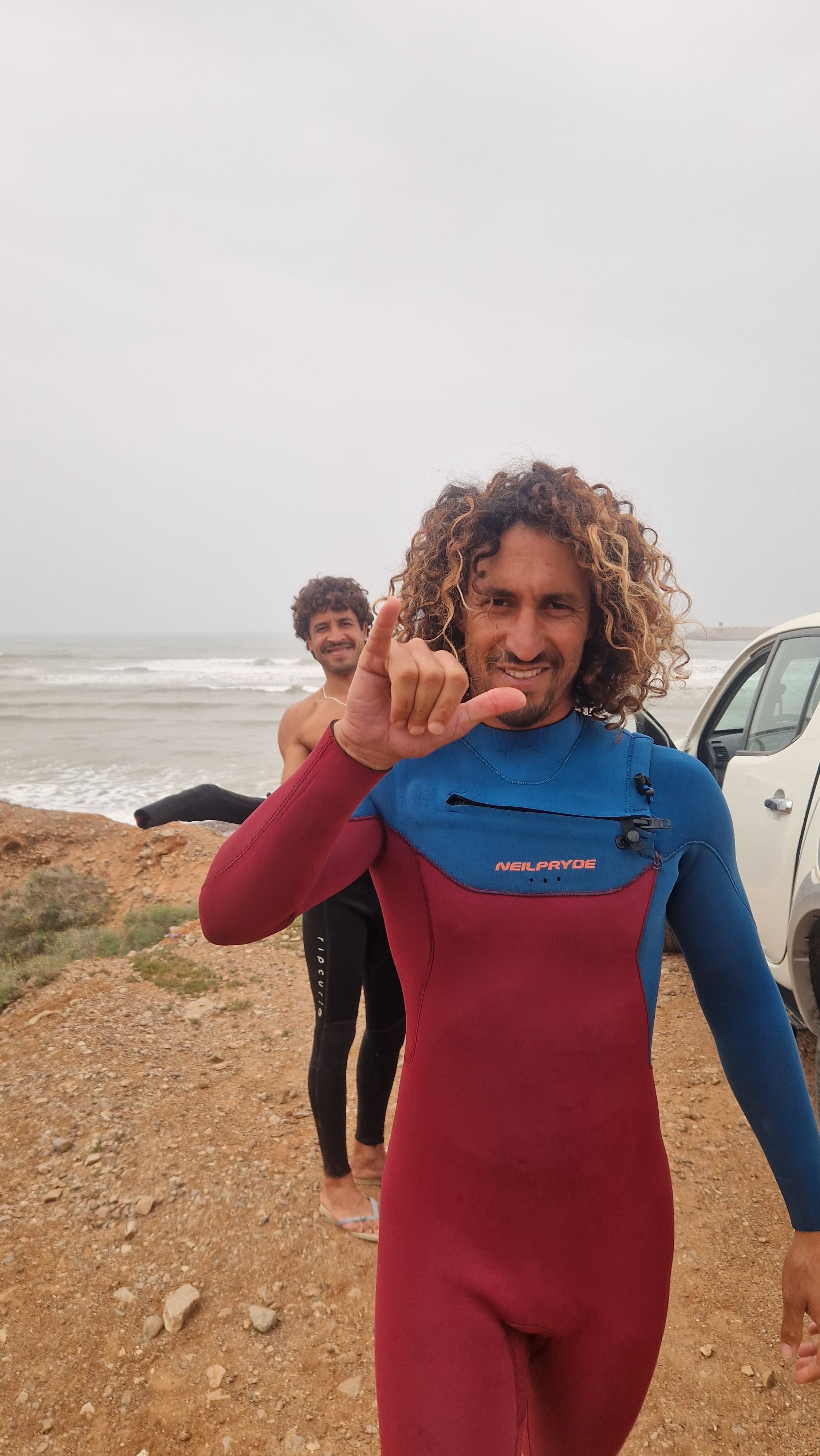 Moha from HeyOla. Two men in wetsuits near a beach, one making a shaka sign and smiling, cloudy sky in the background.