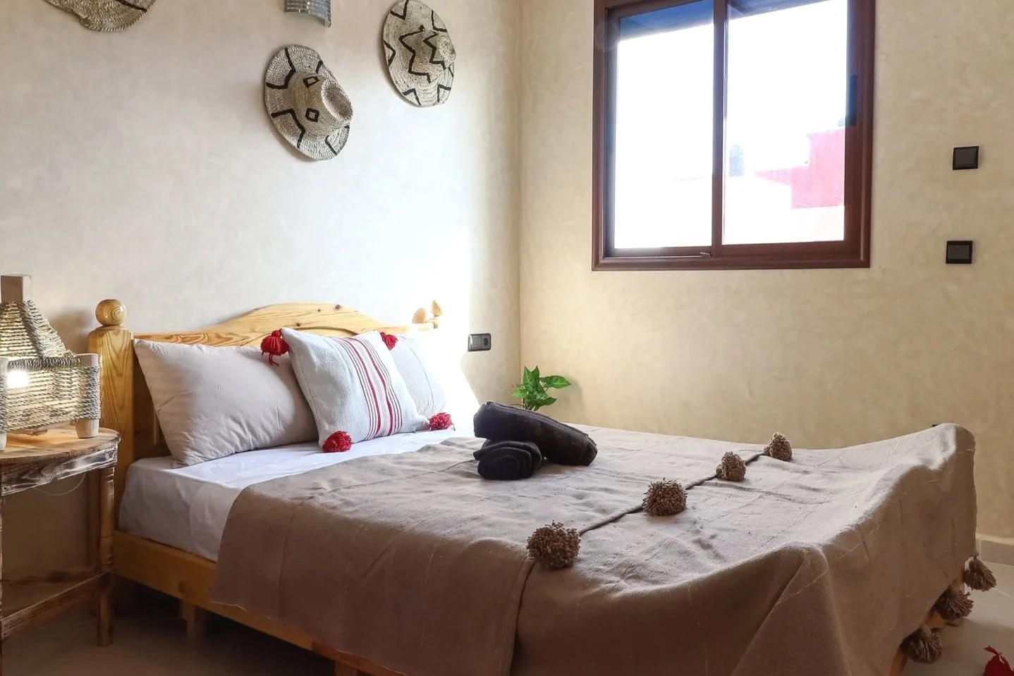 A cozy bedroom with a wooden bed, white pillows, and a beige bedspread decorated with brown pom-poms. Black rolled towels and a decorative pillow with red tassels are on the bed. A side table with a lamp is beside the bed, and a small green plant is 