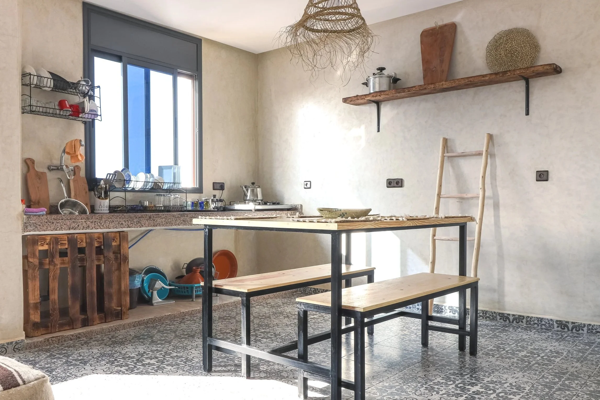 A rustic kitchen with a wooden dining table, benches, and a shelf on the wall decorated with baskets and wood accents, near a window with dishes and kitchenware on the counter.