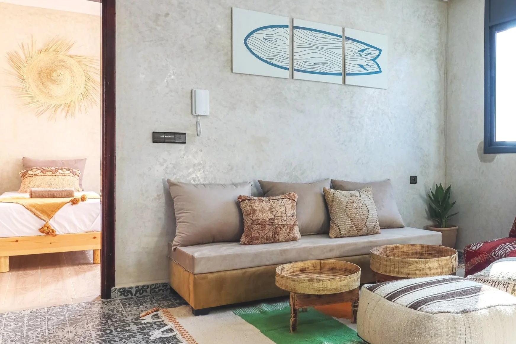Living room with beige suede sofa and three decorative pillows, small wooden coffee tables, potted plant, wall art, window, and a bedroom visible through doorway.