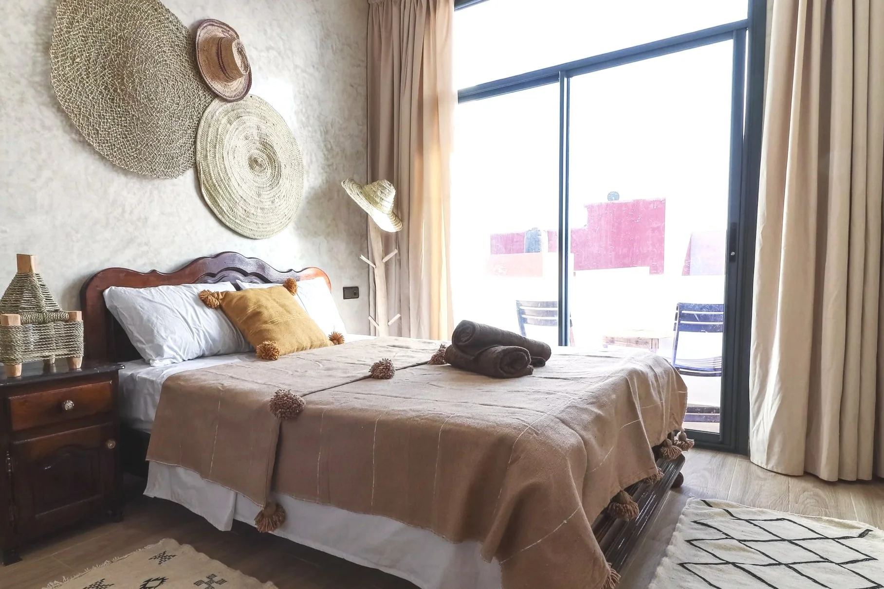 A cozy bedroom with a large bed covered in beige bedding and tassel decorations, a yellow throw pillow, and two rolled towels. The wall behind the bed is decorated with woven hats. There is a side table with a woven lamp, a floor lamp with a straw sh