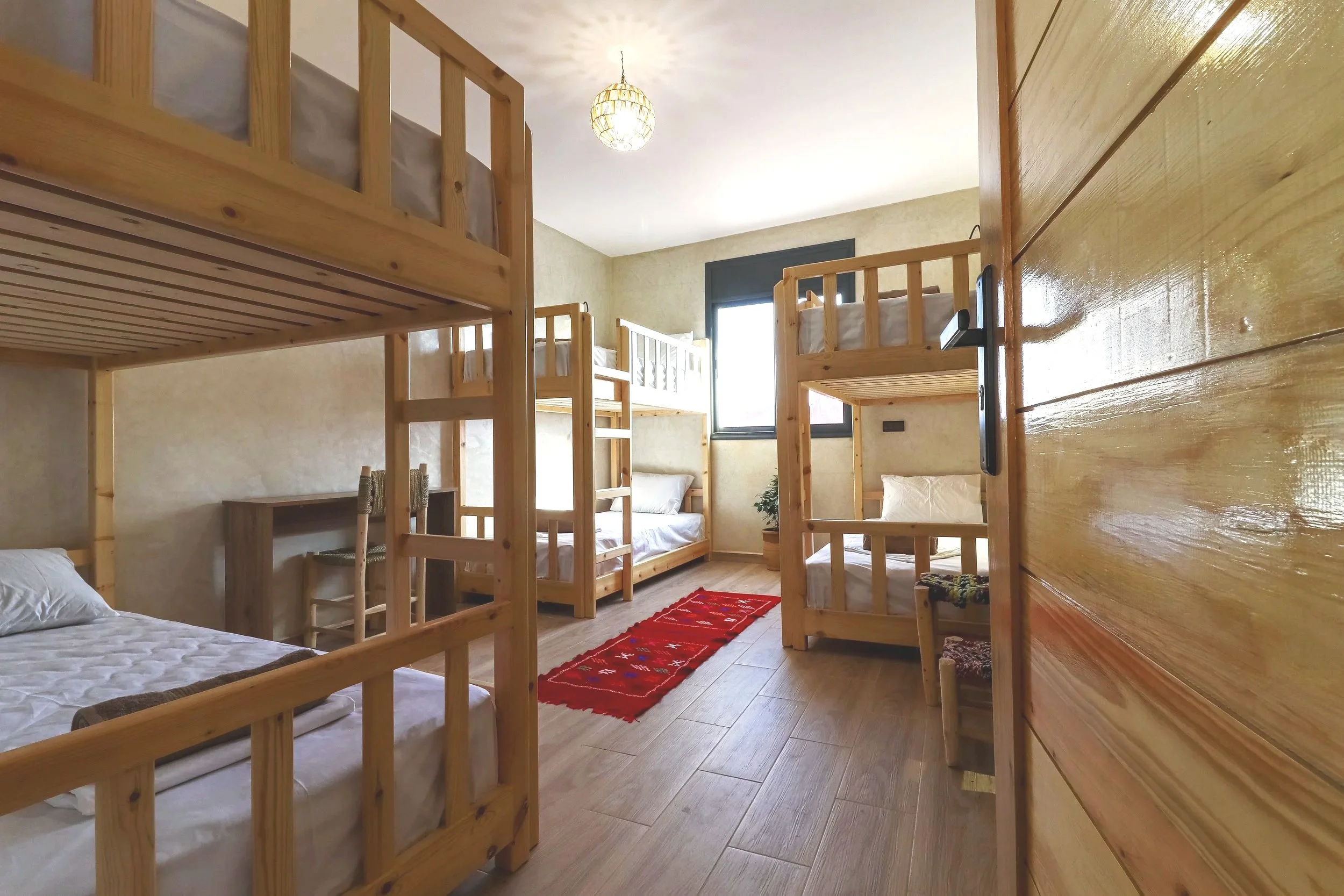 A room with multiple wooden bunk beds, a small desk with a chair, a window with black trim, a red rug on the wooden floor, and a potted plant near the window.