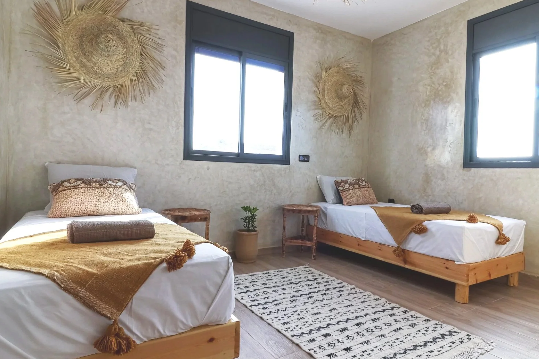 A bedroom with two single beds with white bedding, tan blankets with tassels, and pillows. A small wooden side table with a potted plant sits between the beds. The wall has two woven straw wall hangings, and there are two windows with black frames le