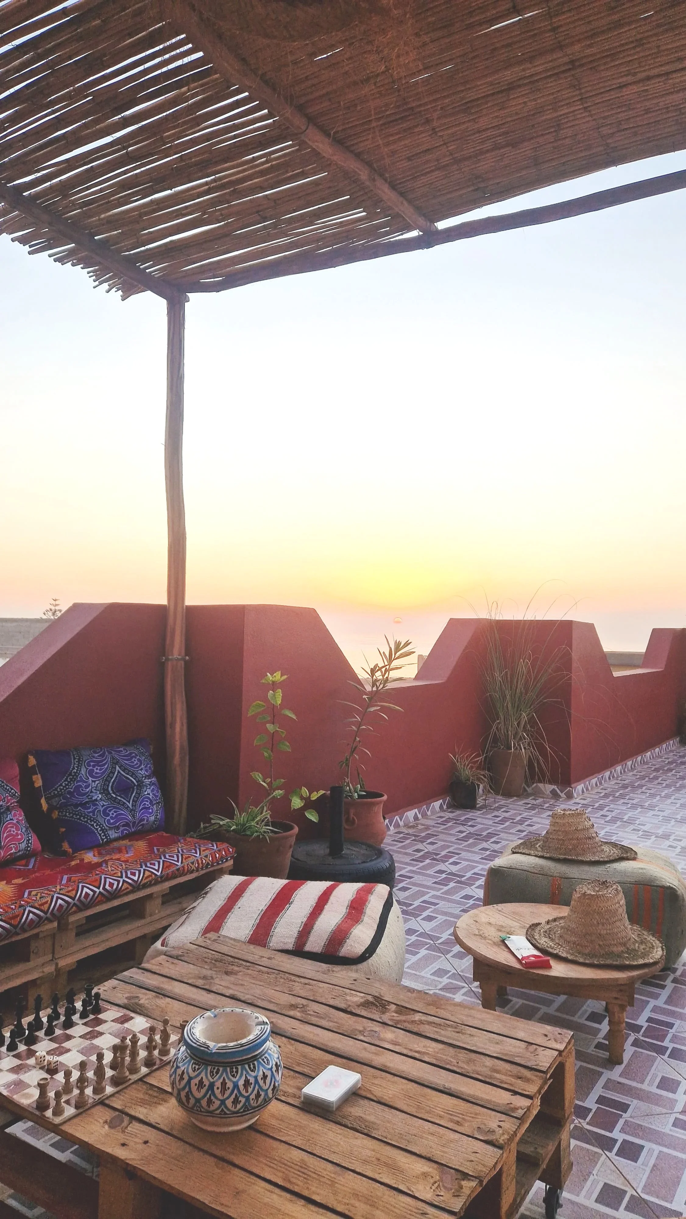 A rustic terrace with potted plants, straw hats, and wooden furniture, overlooking a sunset.