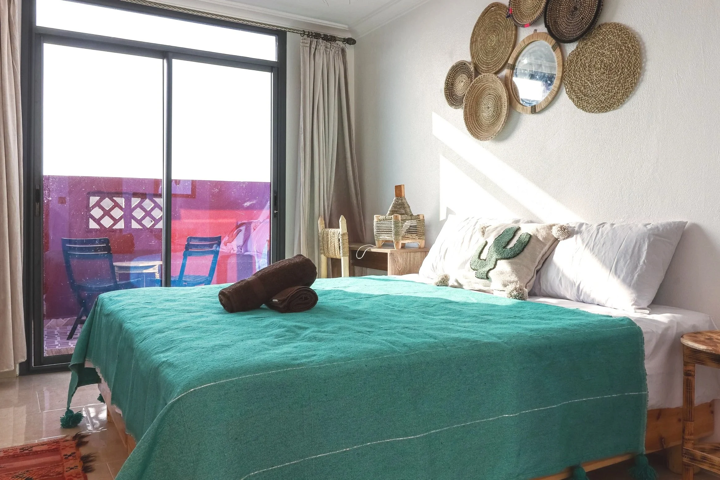 Sunlit bedroom with a bed covered in a turquoise blanket, pillows, and rolled towels, next to a sliding glass door leading to a balcony with chairs, and decorated with woven wall art and a cactus-themed pillow.