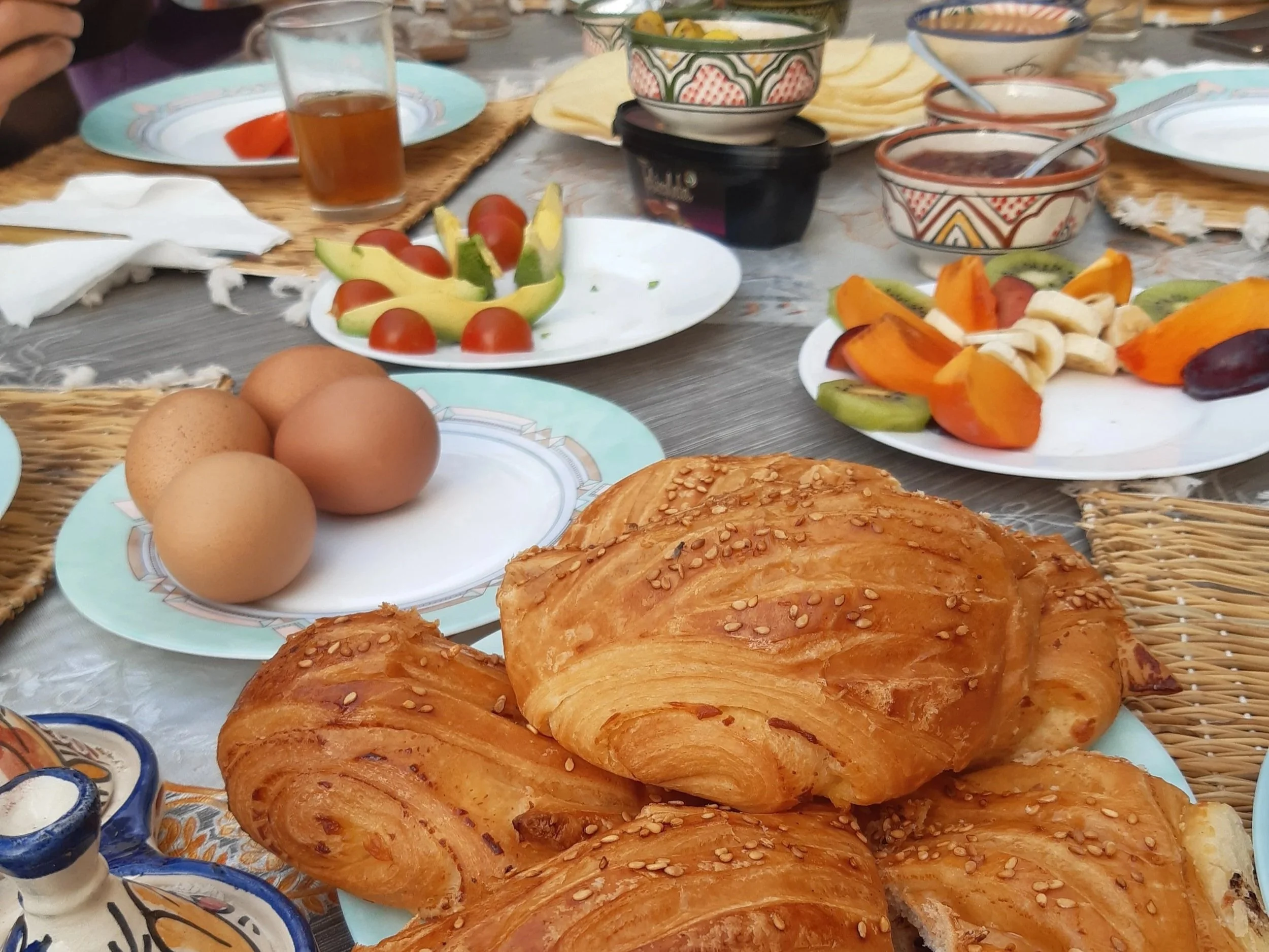 A breakfast table with croissants, eggs, fresh fruit, cherry tomatoes, avocado slices, and various bowls with dips.