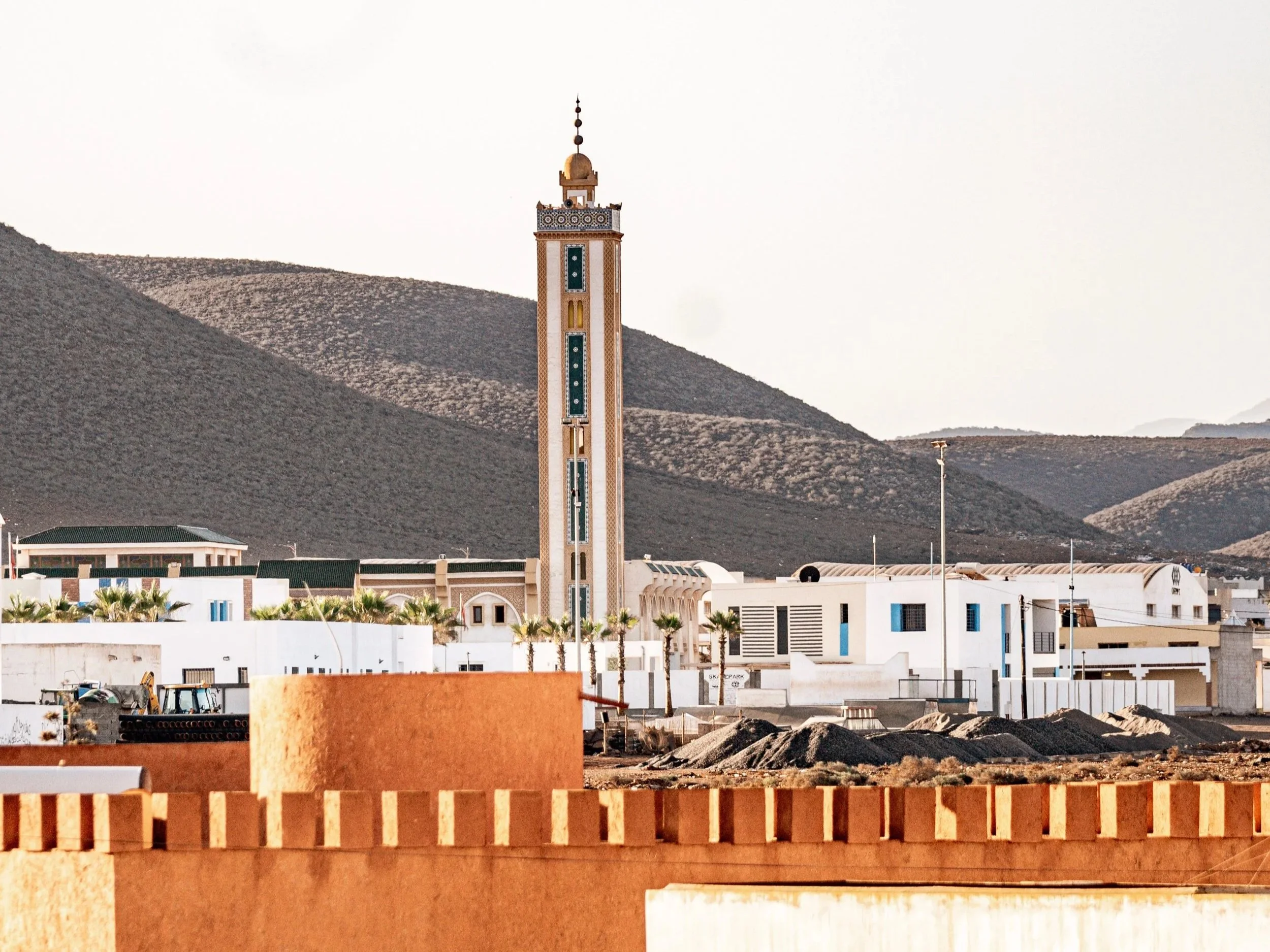 A desert town with white buildings, a tall minaret with decorative tiles, mountains in the background, and construction materials in the foreground.