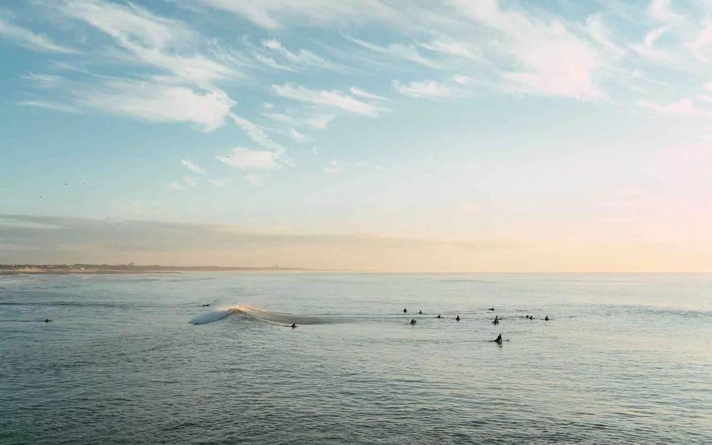 Ocean view with surfers riding gentle waves during sunset, with Soft clouds and distant shoreline visible.