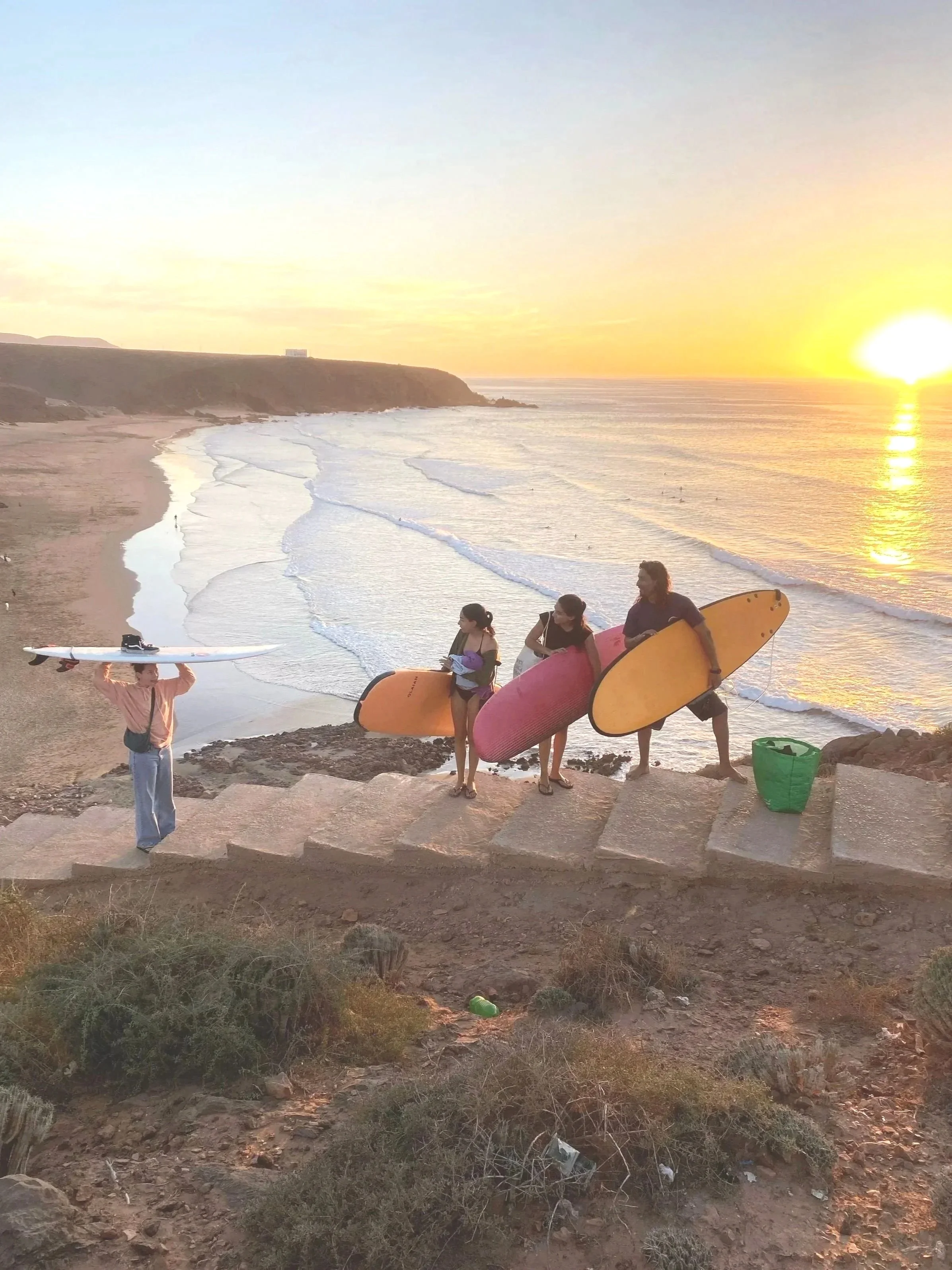 Four people, three holding surfboards, at a beach (Tamhrouchte) during sunset, with another person carrying a surfboard on their head, and a green bag on the ground.