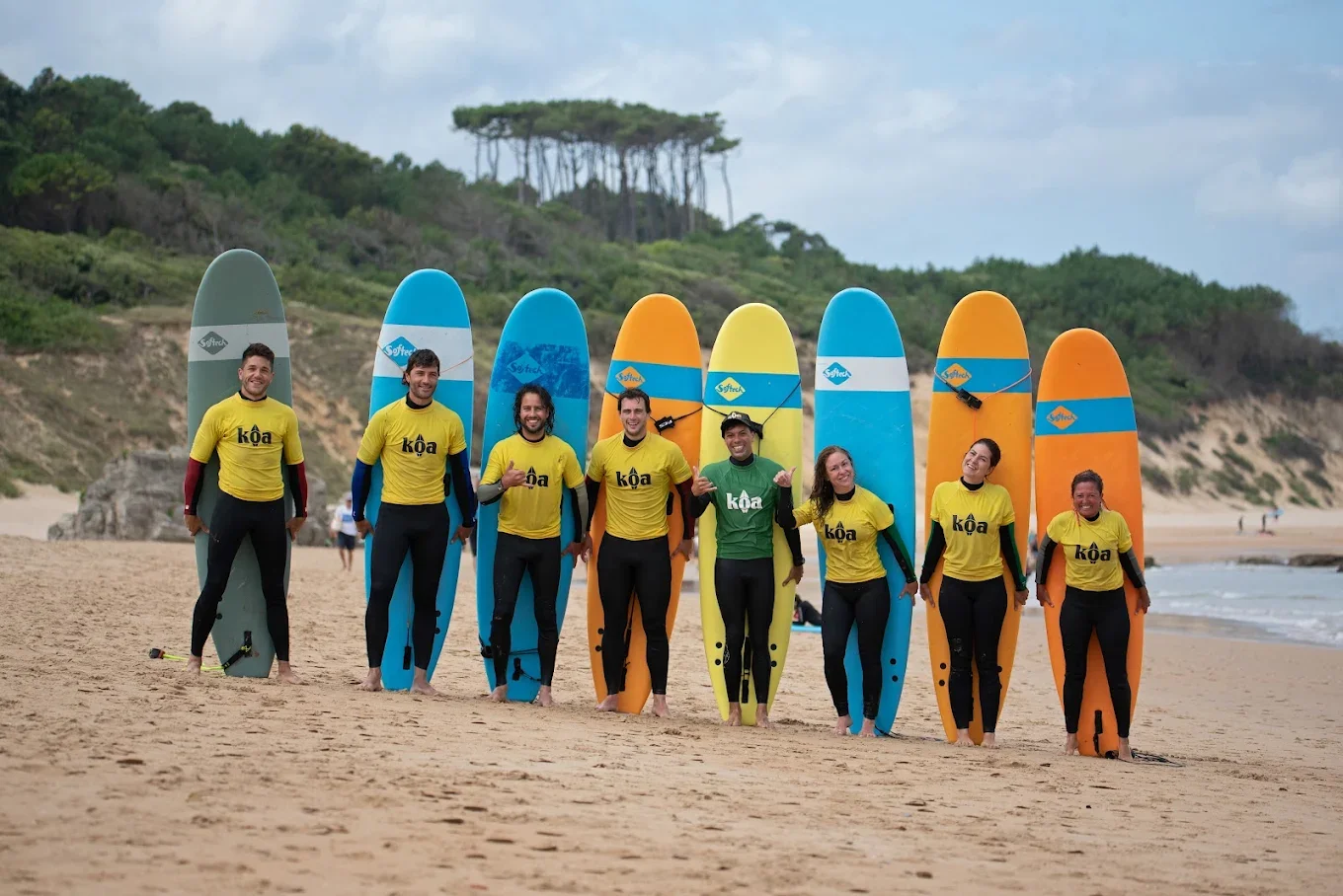 Group of eight people standing on a beach, holding paddleboards, dressed in wetsuits and yellow team shirts with the word 'Koa' on them, with a backdrop of greenery and a cloudy sky.