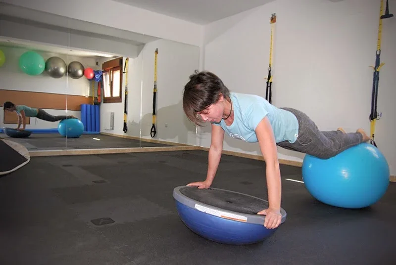 Woman performing an exercise on a balance trainer in a gym, with exercise balls and resistance straps in the background.