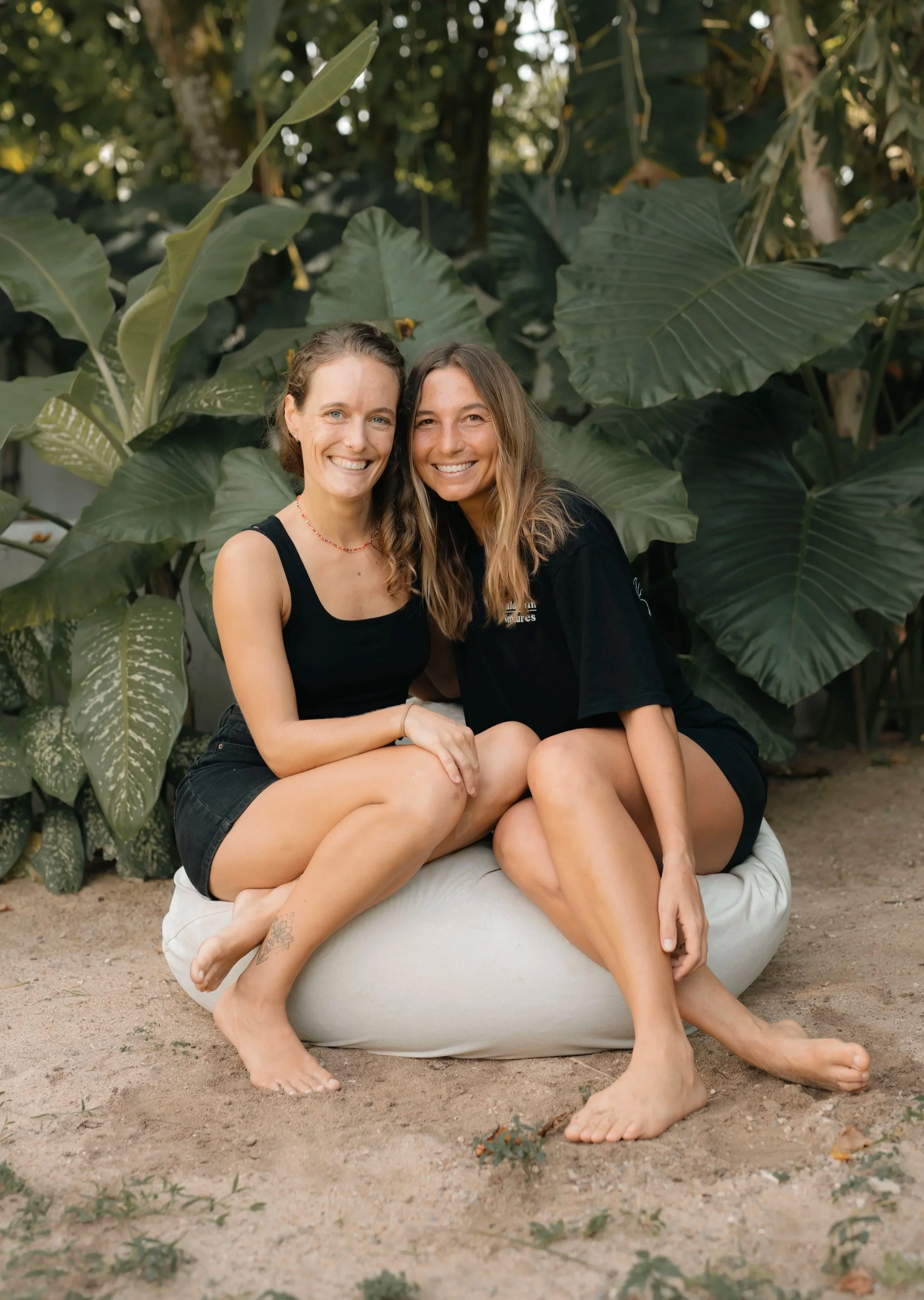 Anni and Daisy, two women sitting closely together on a bean bag outdoors, surrounded by large green tropical leaves, smiling at the camera.