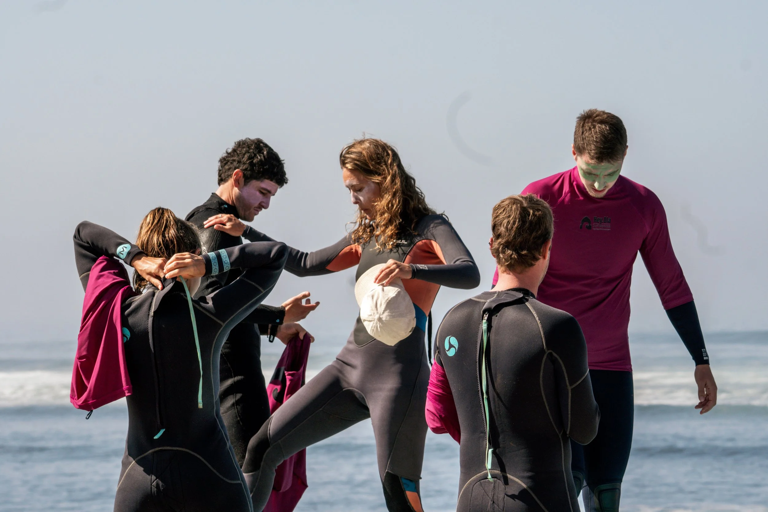 Group of five people in wetsuits standing on the beach, some with backpacks, with ocean waves in the background.