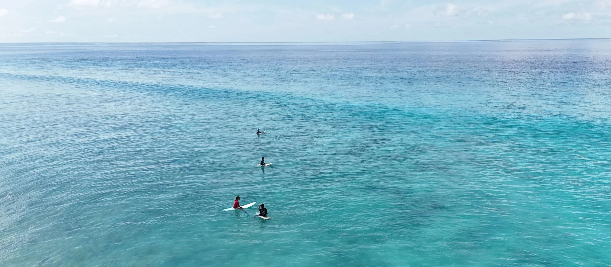 Four surfers sitting on their surfboards in the ocean, with a clear horizon and blue sky in the background.