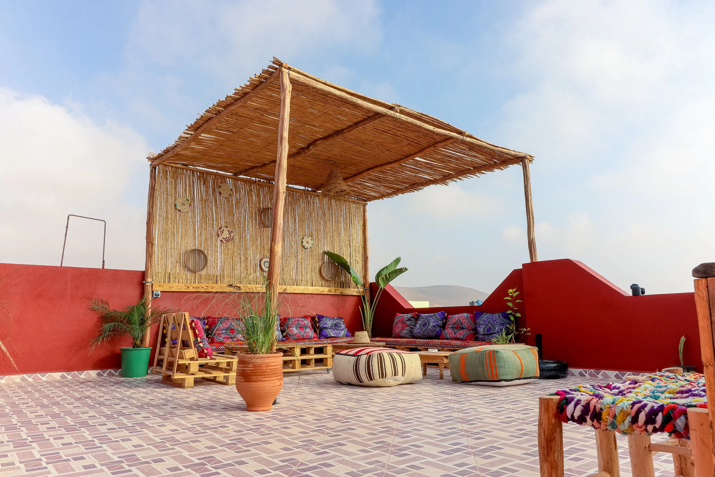 Outdoor seating area with colorful cushions and poufs, potted plants, and a bamboo canopy on a tiled terrace against a blue sky.