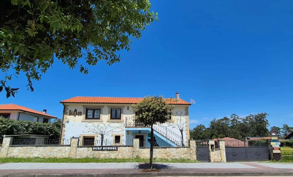 A two-story building with a red-tiled roof, colorful murals, and a small tree in front, situated behind a stone fence with a sign that reads 'Surf School'.