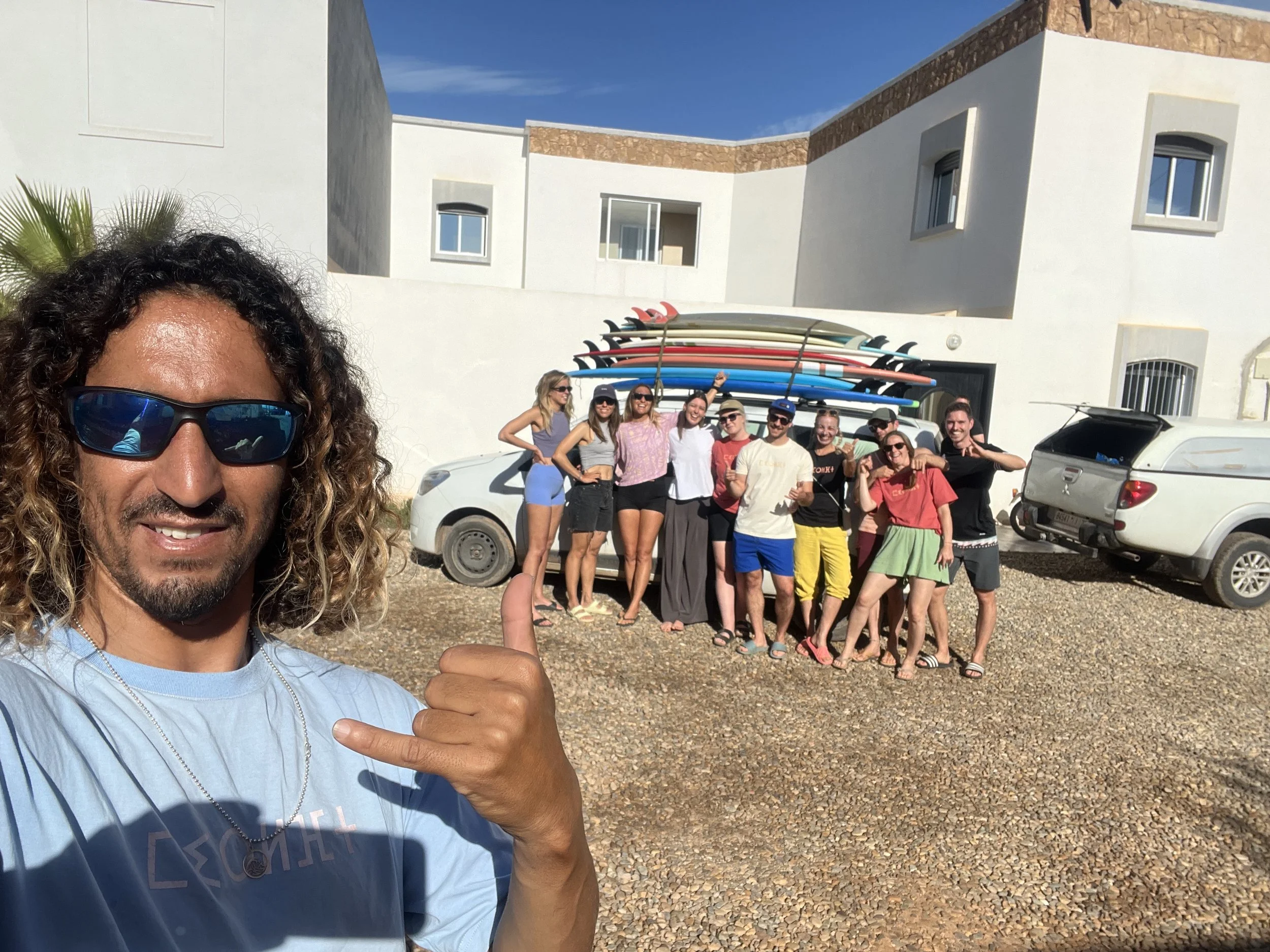 A man in the foreground taking a selfie, smiling and making a shaka sign, with a group of people behind him holding surfboards on a lift, in front of a white building with small windows and a cloudy sky.