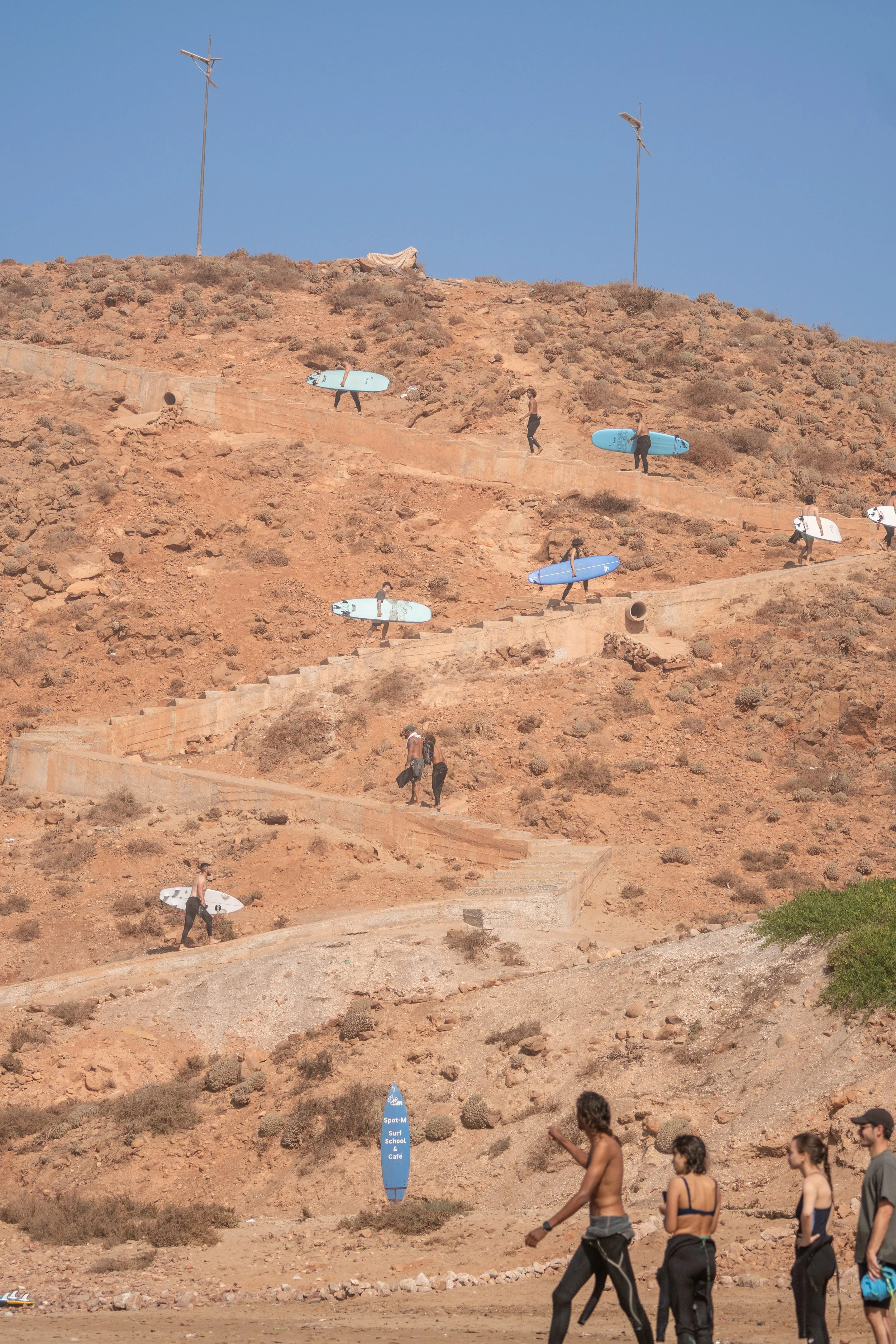 People walking along a sandy beach slope with several individuals carrying surfboards up the hill. In the background, there are two antennas and a blue flag that reads 'Spot M Surf School & Cafe'.