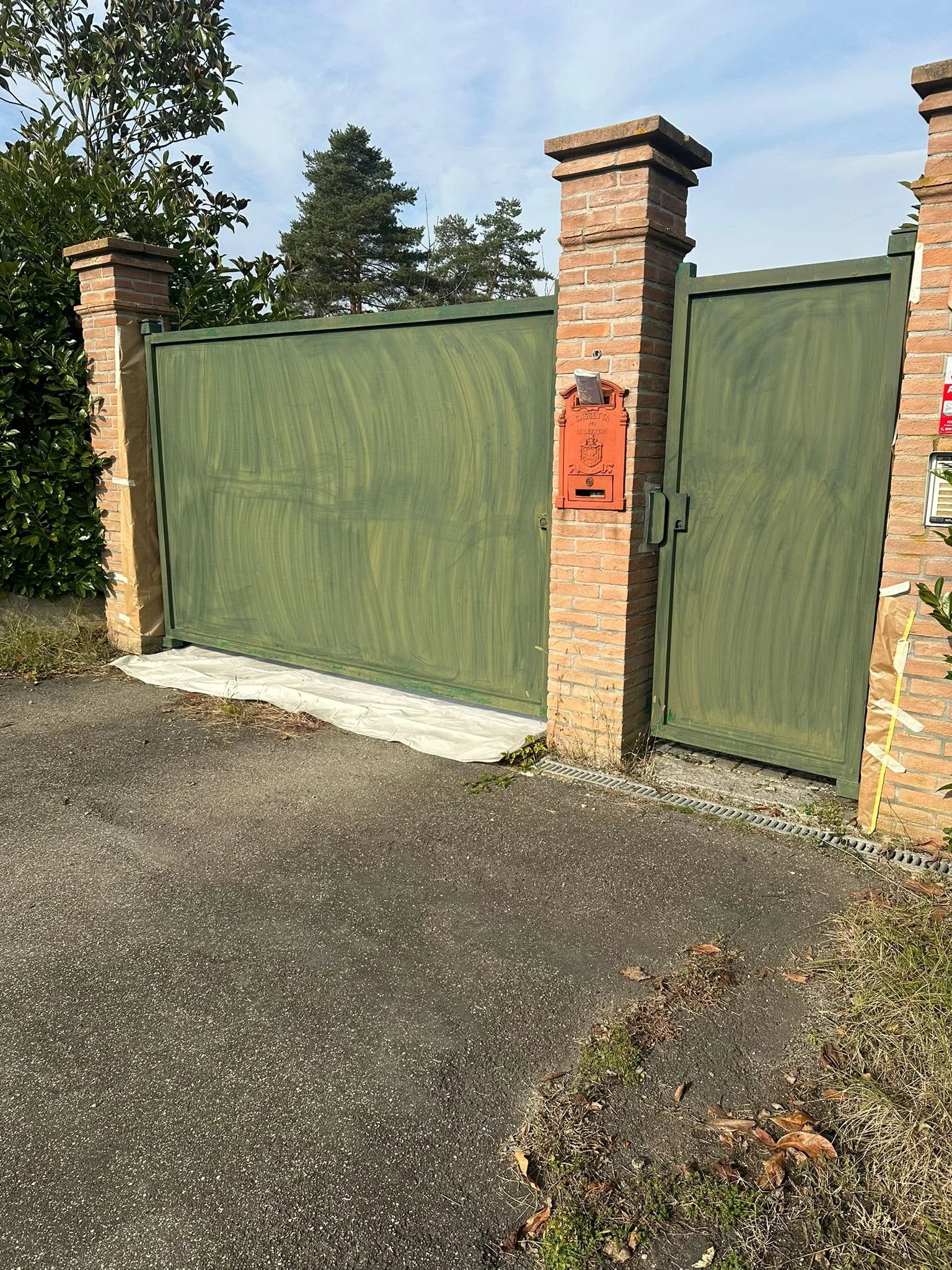 Cancello e portina verde con pali di mattoni, scatola postale rossa e un alberello vicino, strada asfaltata e cielo sereno.