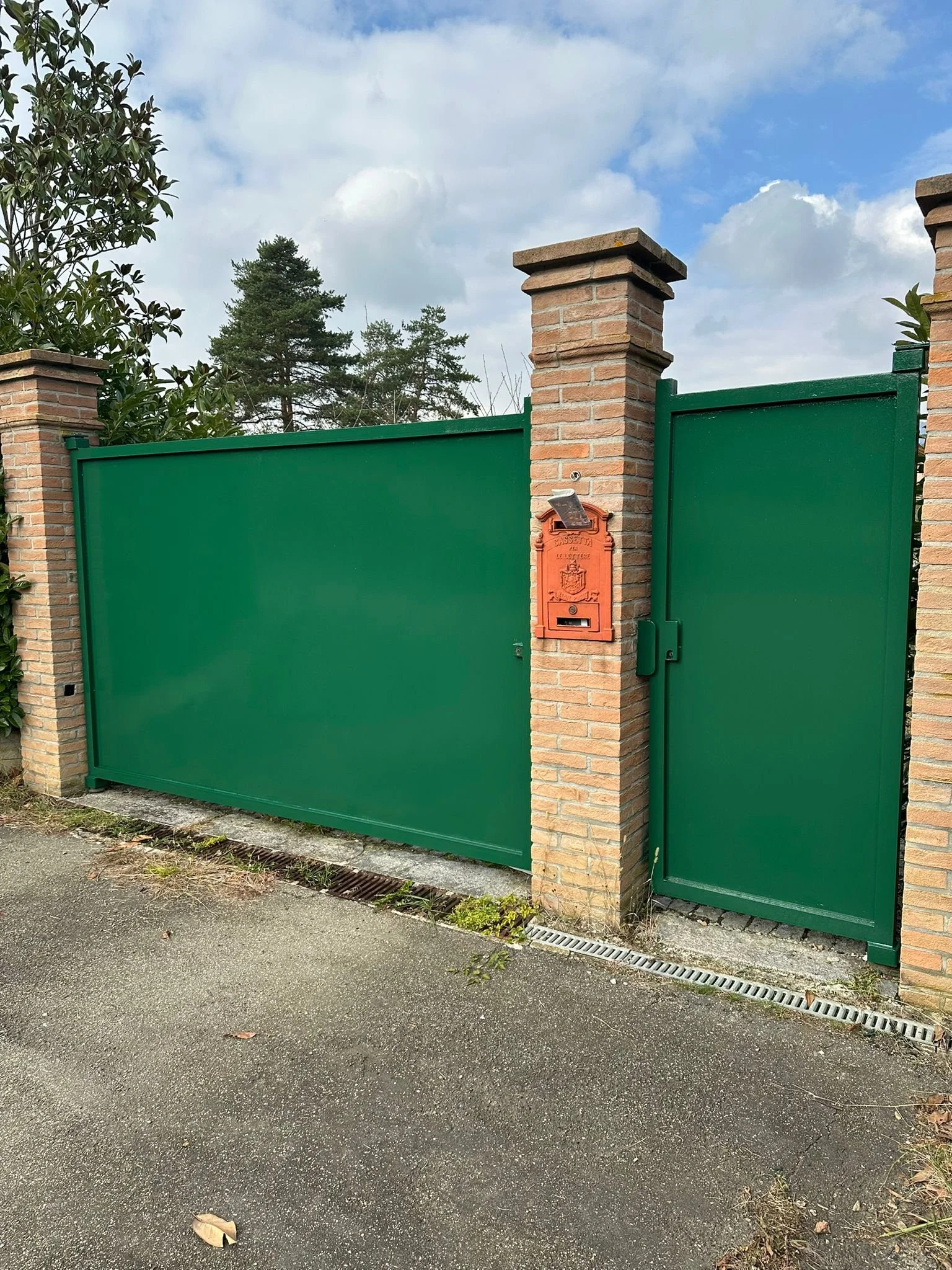 Cancello verde con pilastri di mattoni e cassetta postale vicino a una strada di asfalto, con alberi sullo sfondo e cielo nuvoloso.